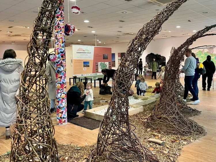 Indoor exhibit with large woven branch sculptures resembling roots or tree trunks, surrounded by children and adults engaging with educational displays about Charnwood Forest.