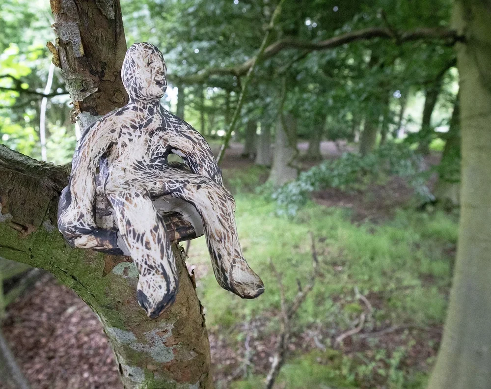 A sculpture of a sitting human figure with a pattern resembling tiger stripes, mounted on a tree branch in a wooded area.