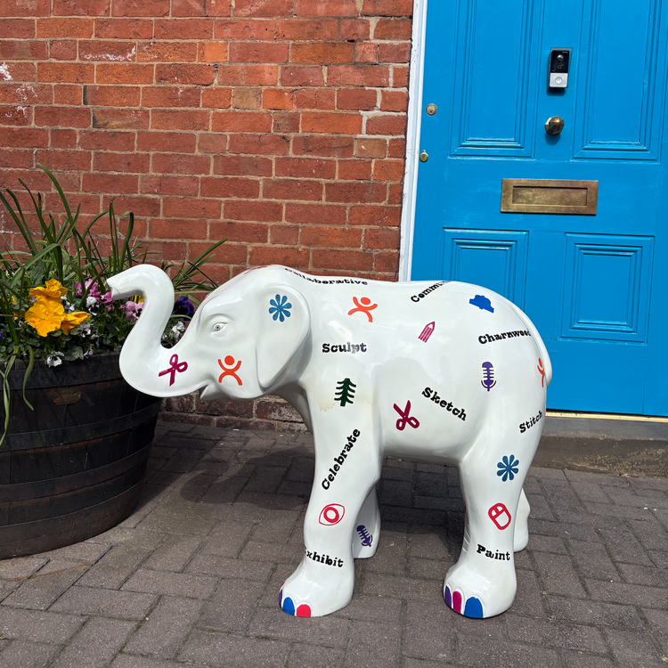 Decorative white elephant sculpture with arts and crafts themed words and symbols, positioned outside near a brick wall and a blue door, beside a large planter with yellow, purple, and white flowers.