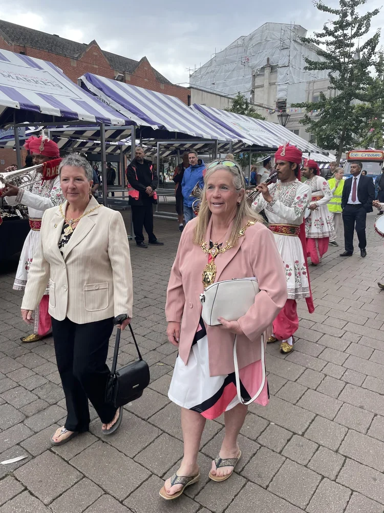 Street scene with people walking past a parade of musicians in traditional costumes playing wind instruments under striped blue and white canopies.