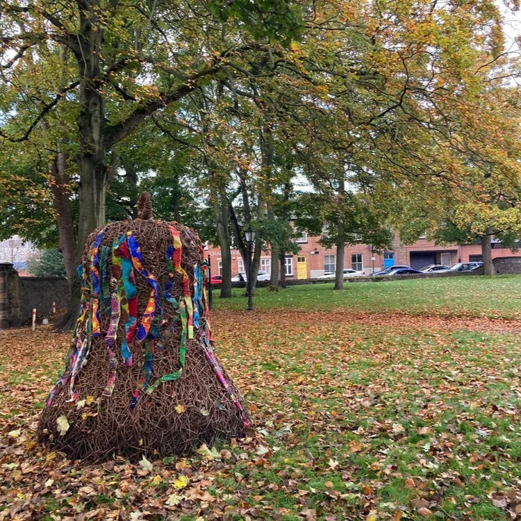 A large, bell-shaped sculpture made of intertwined branches, decorated with colourful ribbons, situated in a park with autumn leaves on the ground and trees with fall foliage as part of the Loughborough Bell Festival 2024.