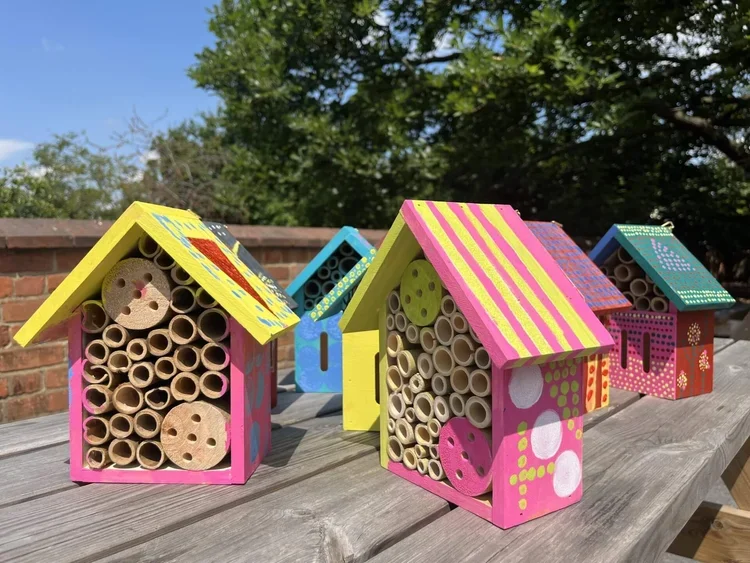 Colorful decorative insect hotels made from painted wood and cardboard tubes, placed outdoors on a wooden surface.