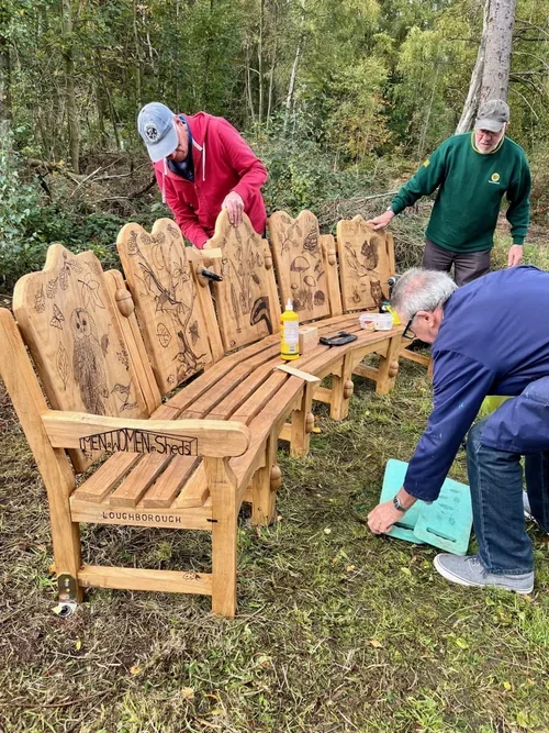 Three men working on a wooden bench with carved and engraved designs in a forested outdoor area.