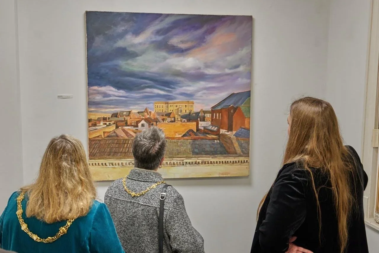 Three people viewing a landscape painting of rooftops and a cloudy sky in an art gallery.