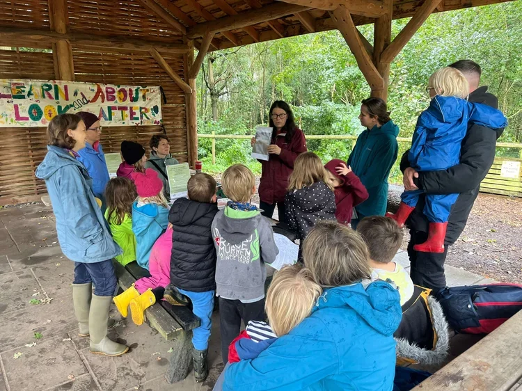 Group of children and adults gathered around a woman reading a paper under a wooden shelter with green trees in the background.