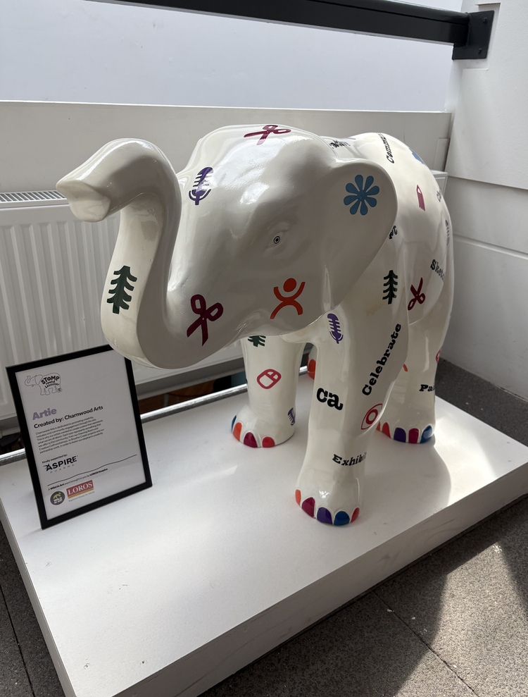 A white elephant sculpture decorated with colorful symbols, words, and logos, displayed on a white platform next to a sign.