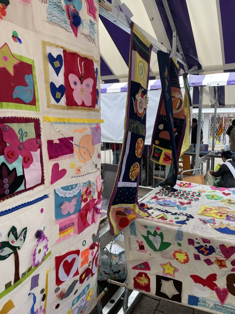 Colorful fabric arts and decorations, including butterflies, hearts, flowers, and various shapes, displayed on tables and hanging on a booth at an outdoor event.
