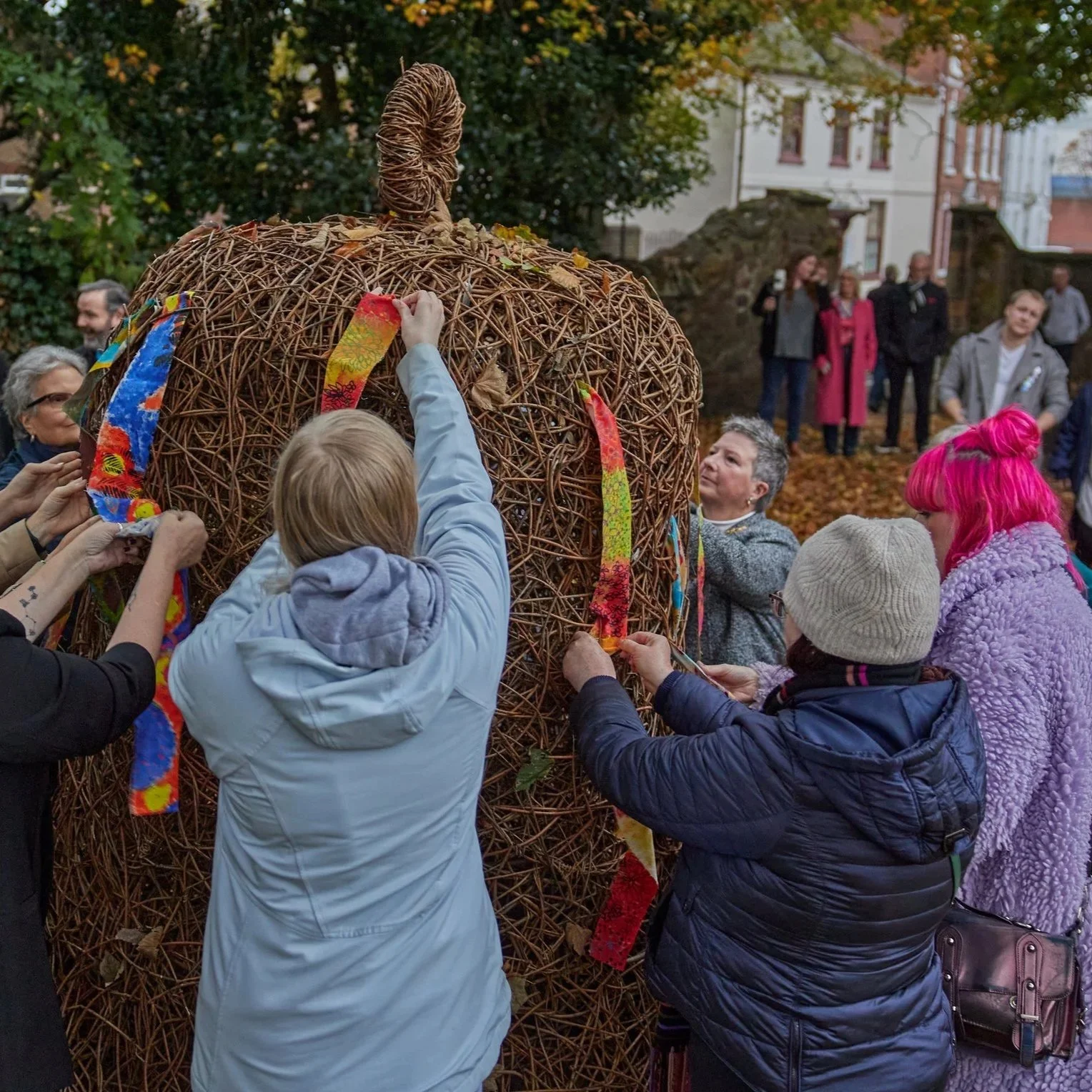 People decorating a large wicker bell as part of the Loughborough Bell Festival with colorful ribbons outdoors.