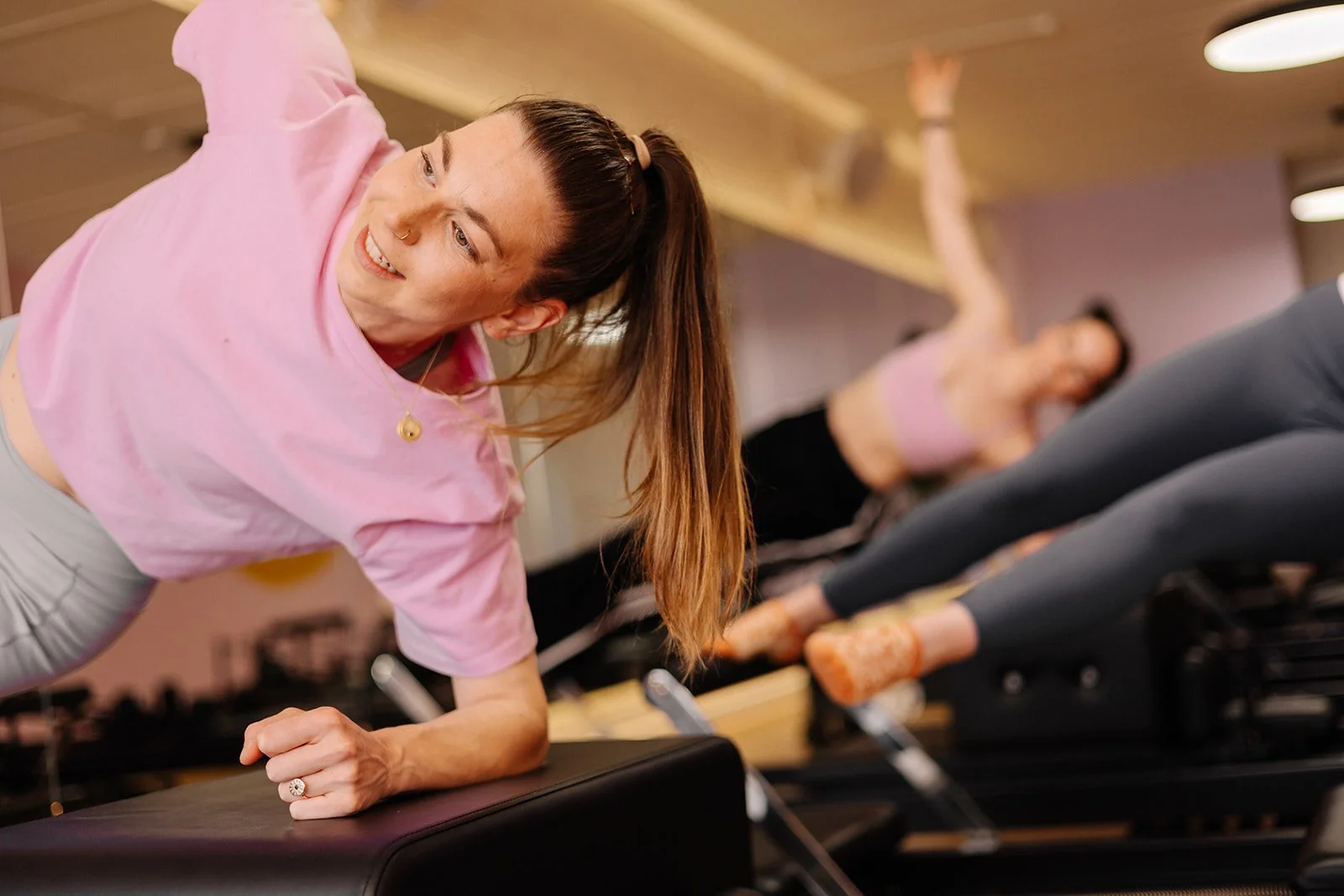 A woman in a pink shirt and gray leggings performing a plank exercise on an elevated platform in a fitness class, smiling and looking towards the camera. Other women are doing the same exercise in the background.