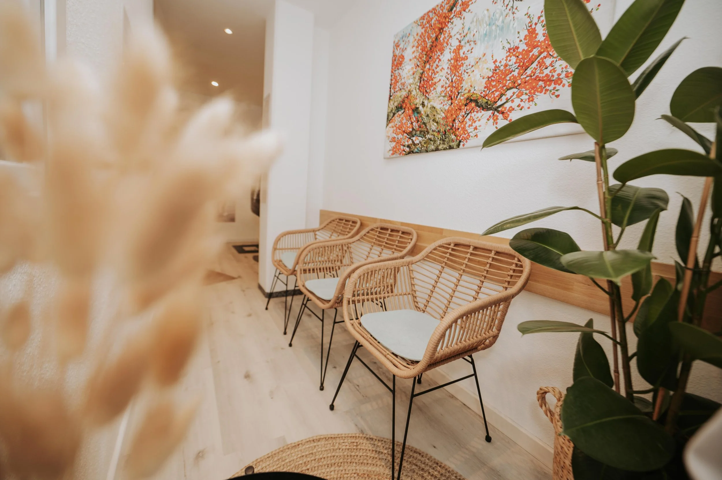 Wartebereich mit drei Rattanstühlen, einer grünen Pflanze und einem Wandbild mit orangefarbenen Blüten auf weißem Hintergrund.
