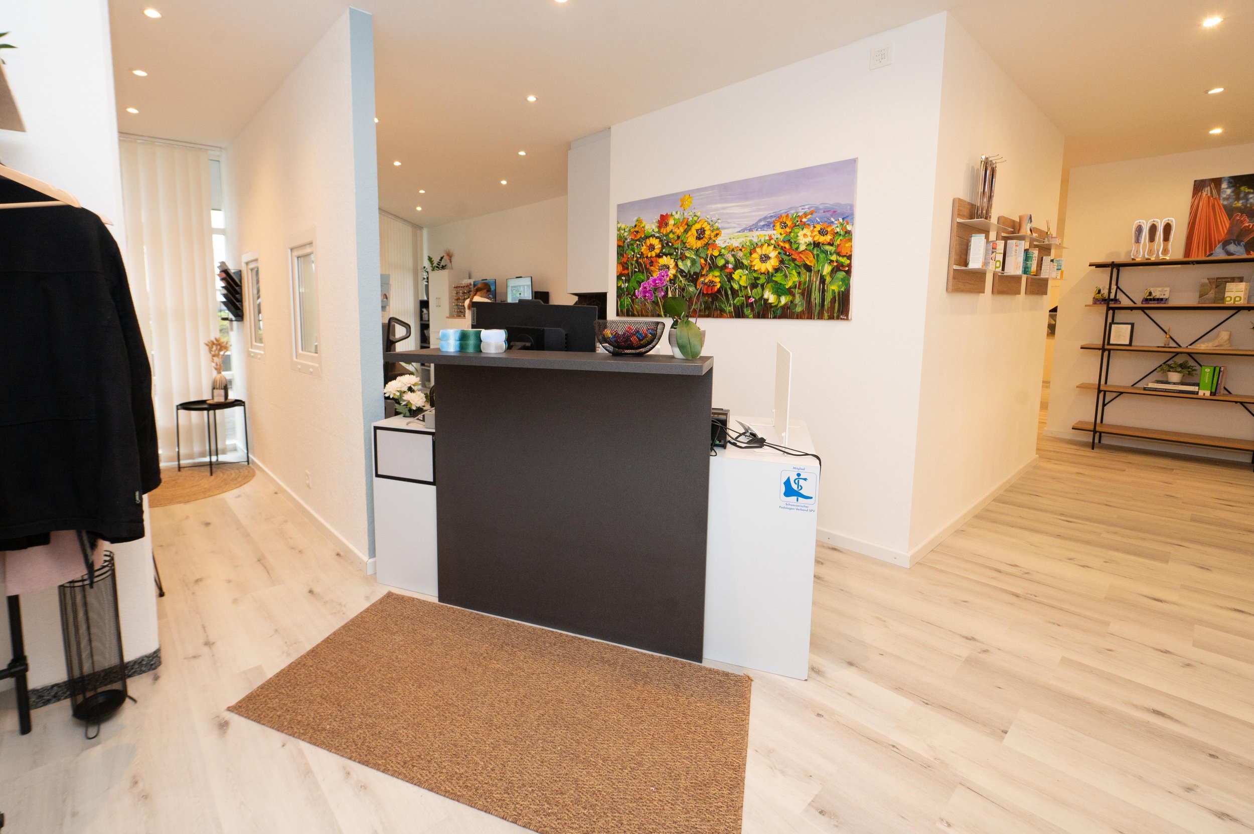 Rezeption in einem modernen Raum mit buntem Blumenbild an der Wand, einem schwarzen Empfangstresen, einer Schale mit Christbaumschmuck, und einem Badezimmer-Handhygiene-Hinweis. Im Hintergrund sind Computer und eine Person sichtbar.