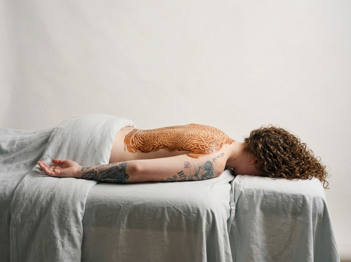 A woman with curly hair and tattoos lying face down on a bed with white sheets and head turned to the side.