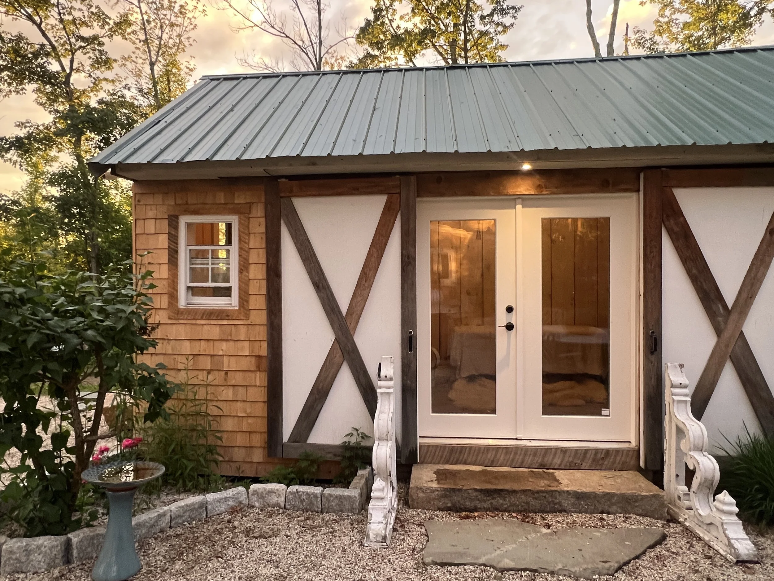 Front view of a small house or cottage with a metal roof, white sliding glass doors, a small window to the left, and decorative wooden and brick siding. There are trees and garden plants around the house, with a birdbath in the front yard and a stone border separating the garden from the gravel area.