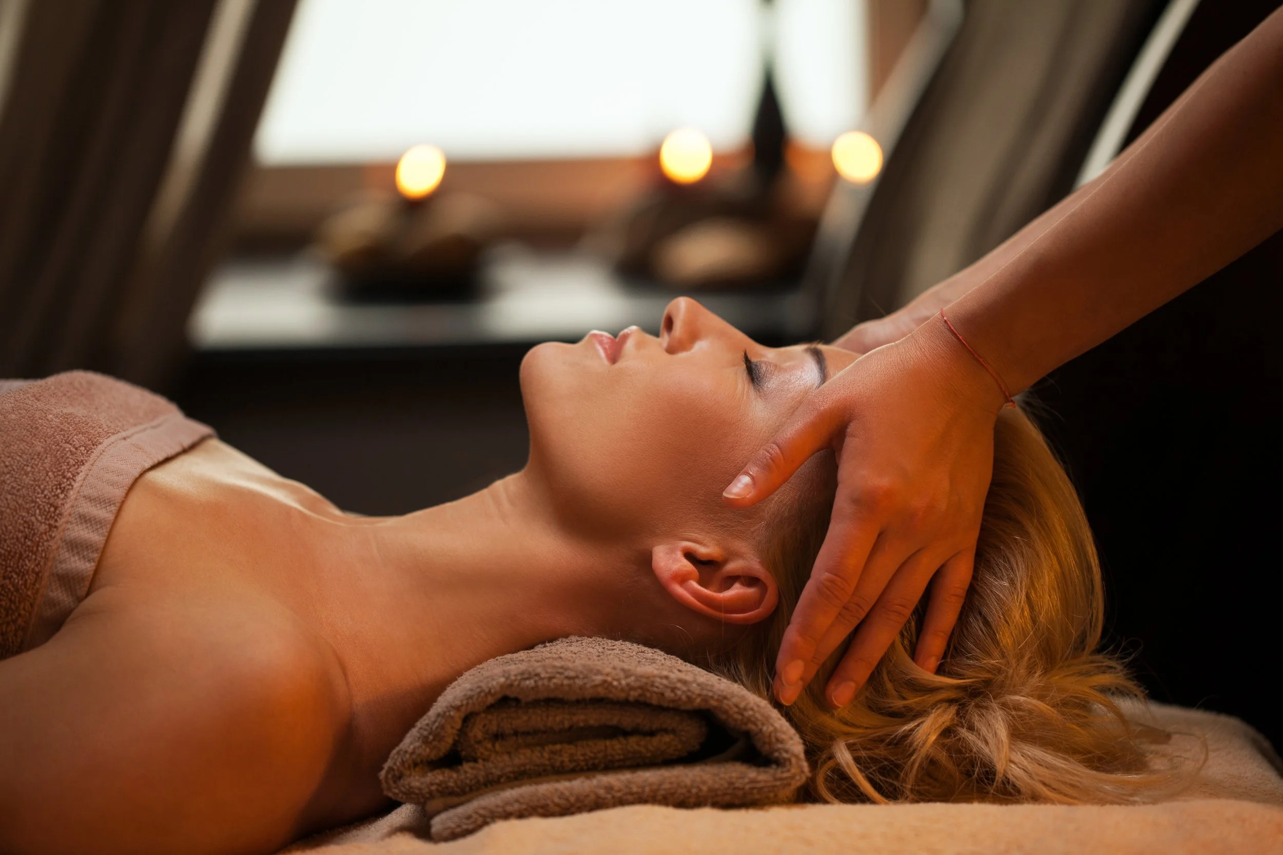 A woman receiving a facial massage at a spa, lying on a treatment table with her head on a towel, with lit candles in the background creating a calming ambiance.