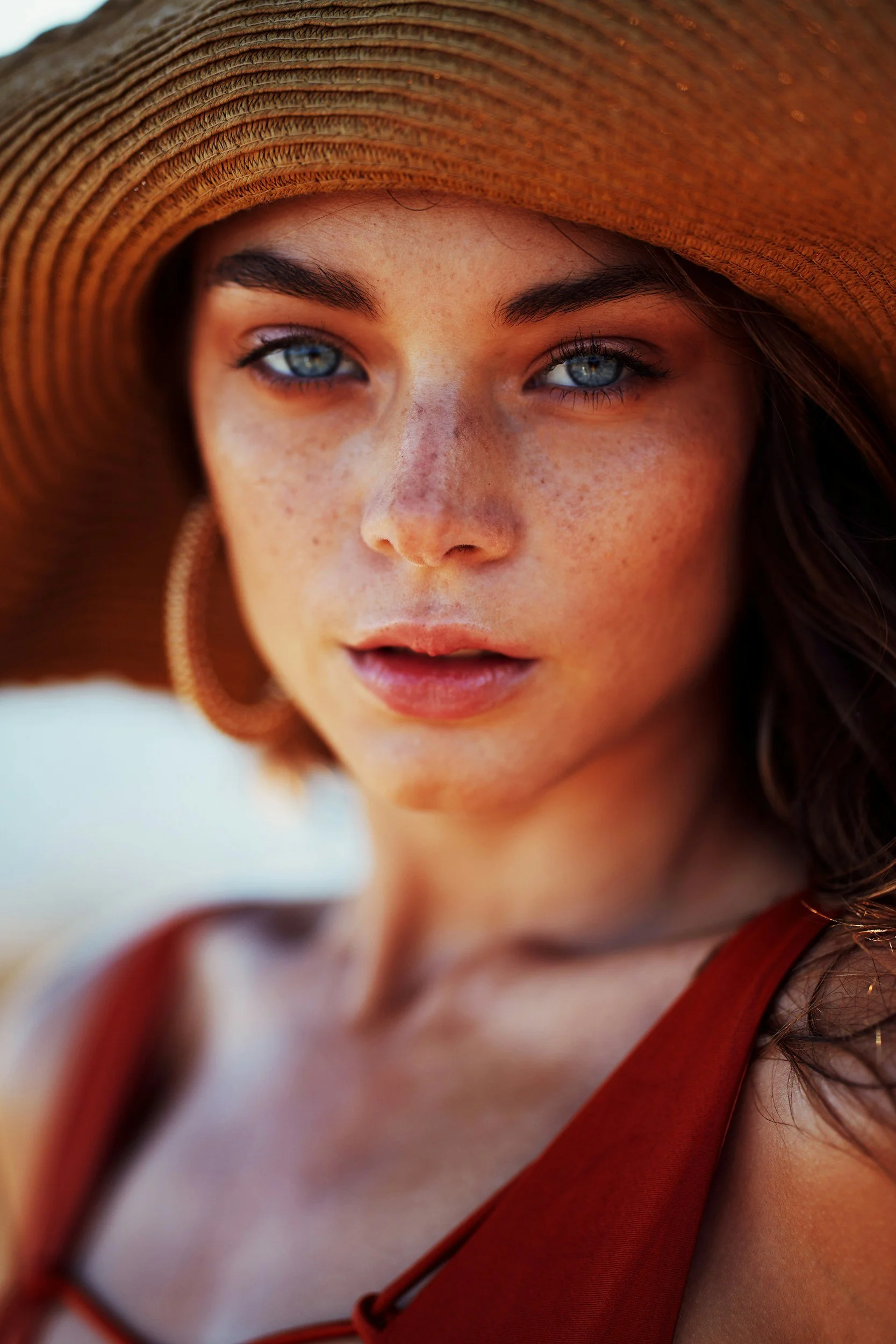 Close-up of a woman with blue eyes, freckles, wearing a large straw hat and an orange dress.