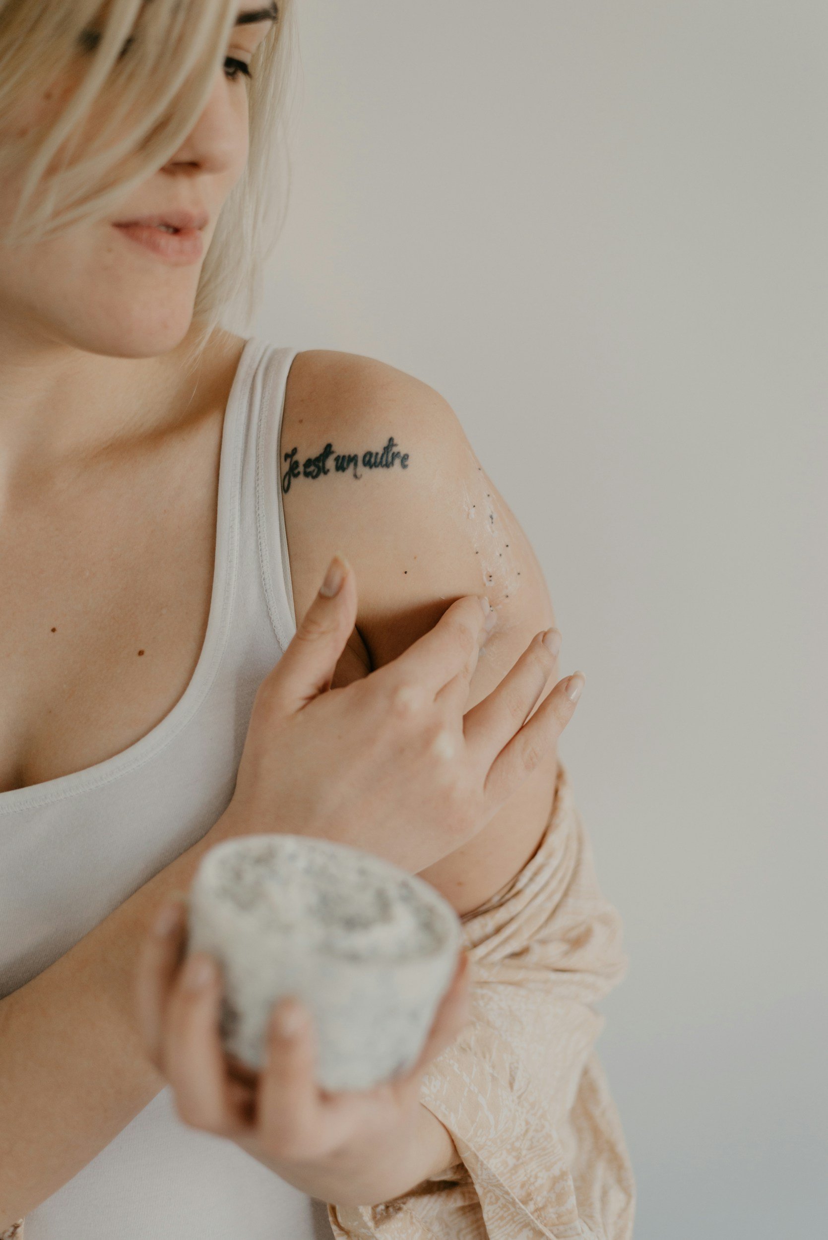 A woman with a tattoo reading 'Je est un autre' on her shoulder, applying lotion to her arm, holding a small bowl in her other hand.