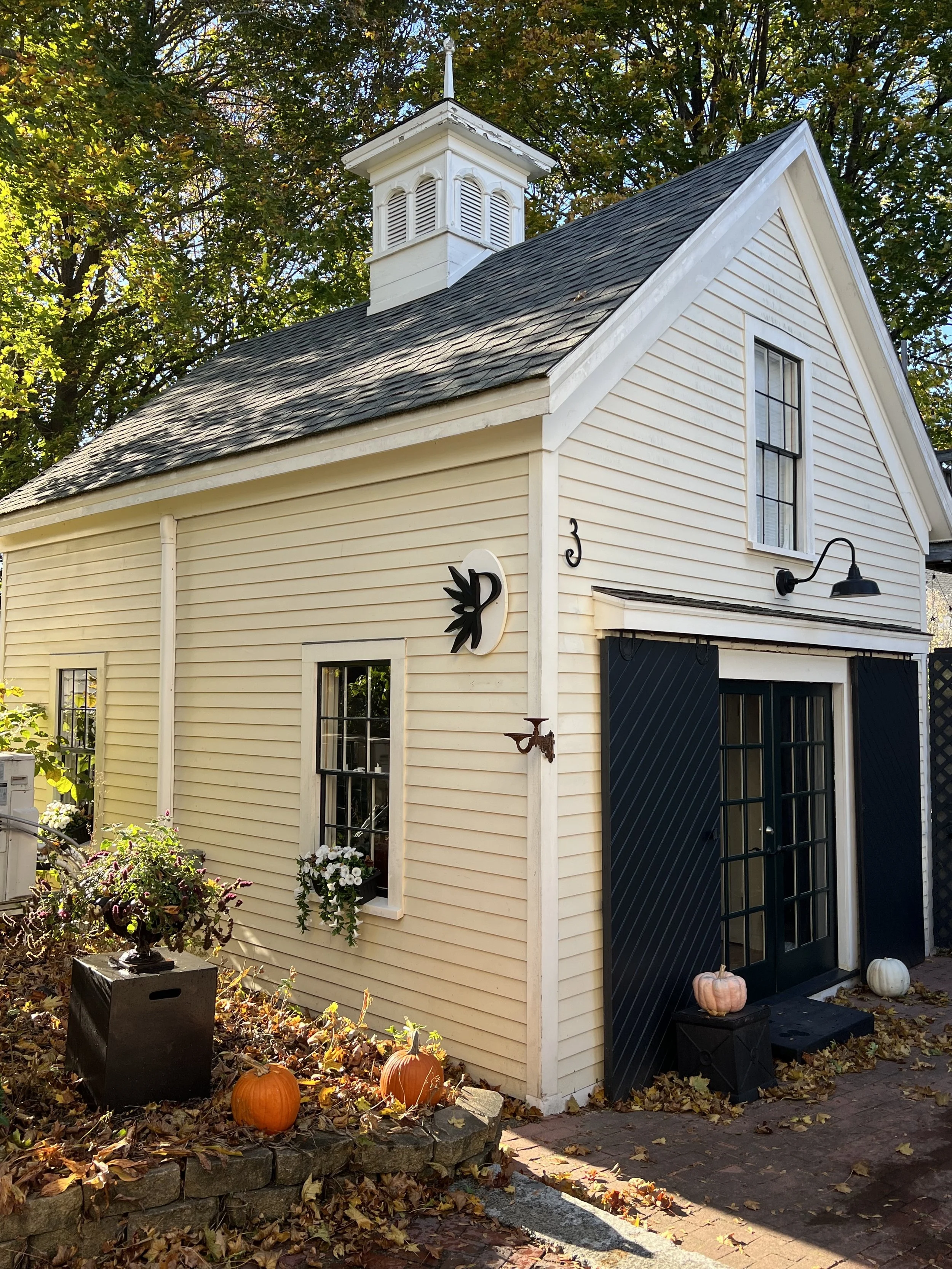 Small cream-colored house with black shutters, pumpkin decorations, and fall leaves outside, surrounded by trees.