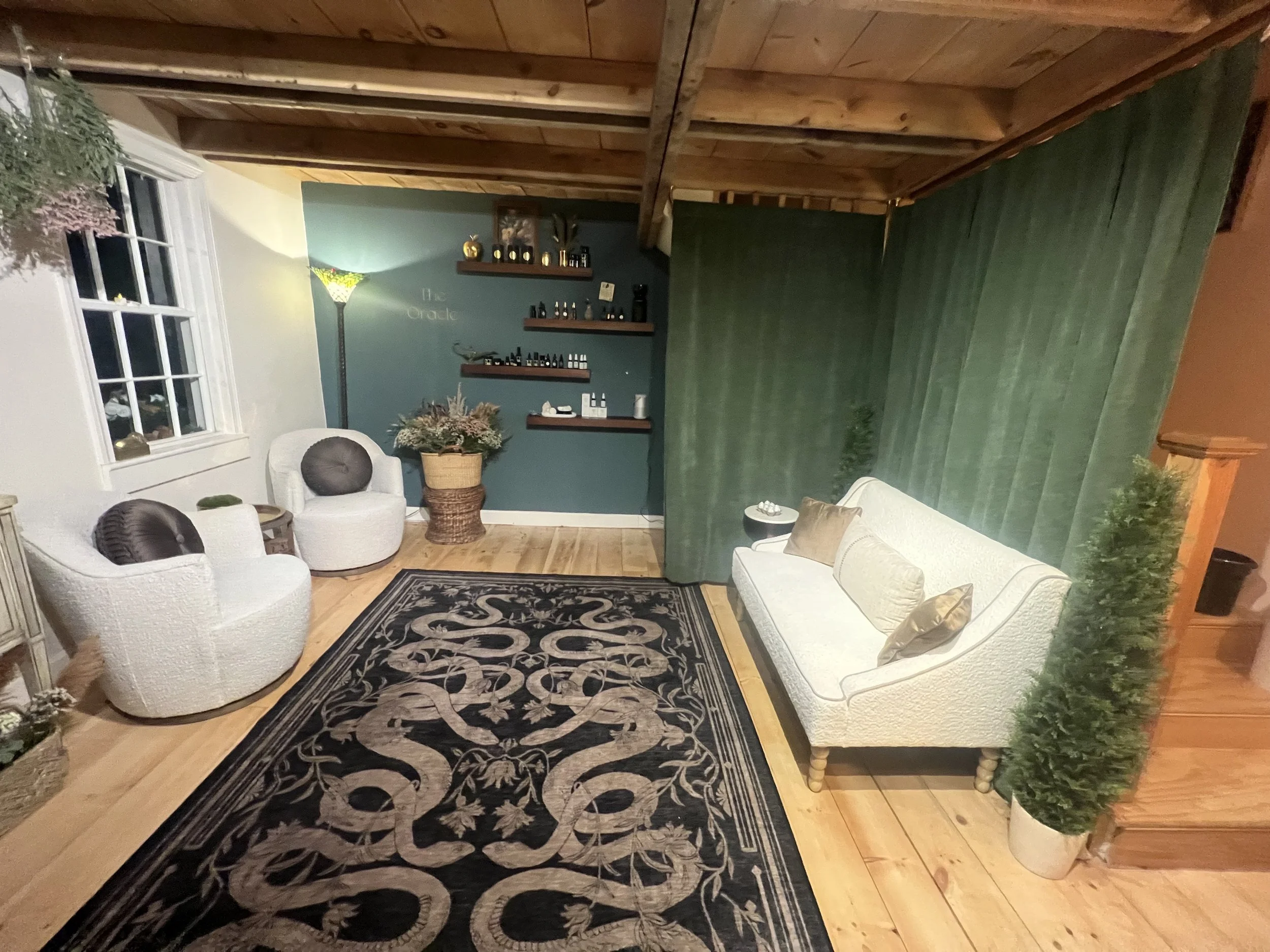 Living room with white textured sofa and two white armchairs, black and beige patterned rug, green curtain wall, teal accent wall with shelves displaying small bottles and decorative items, green potted plants, floor lamp, window with white trim, woo