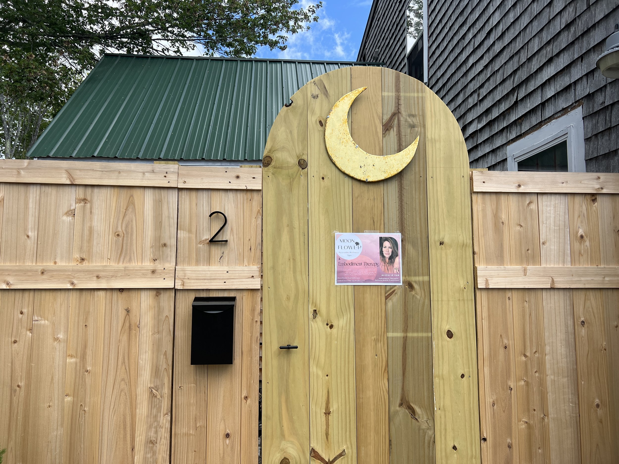 A wooden fence with a yellow, crescent moon decoration on the right side. A poster advertising a therapy session is attached to the fence. The background shows a building with gray shingles and a green metal roof, trees, and a blue sky with some clouds.