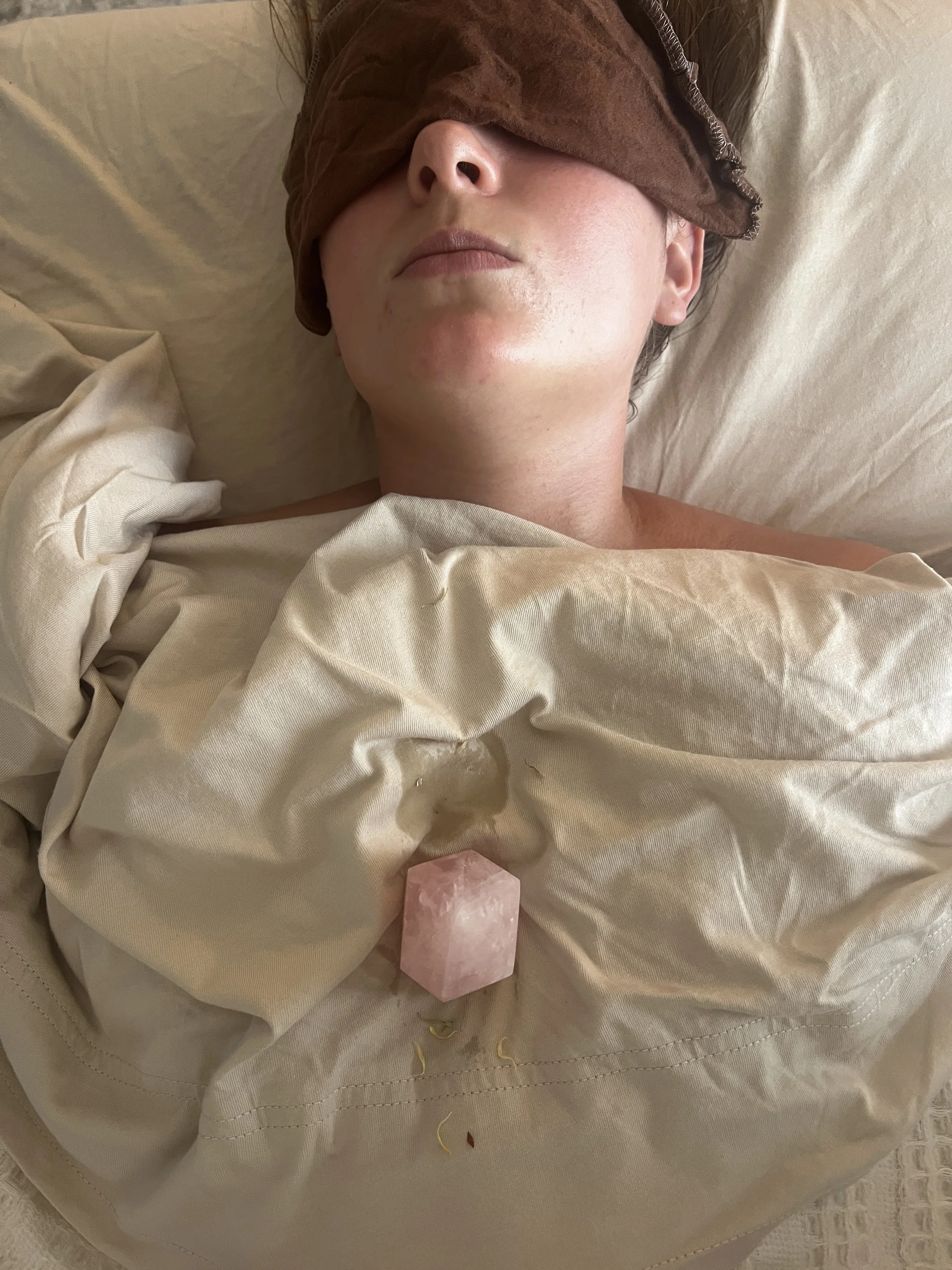 Person lying in bed with a brown sleep mask covering their eyes, soft beige sheets, and a pink salt lamp nearby.