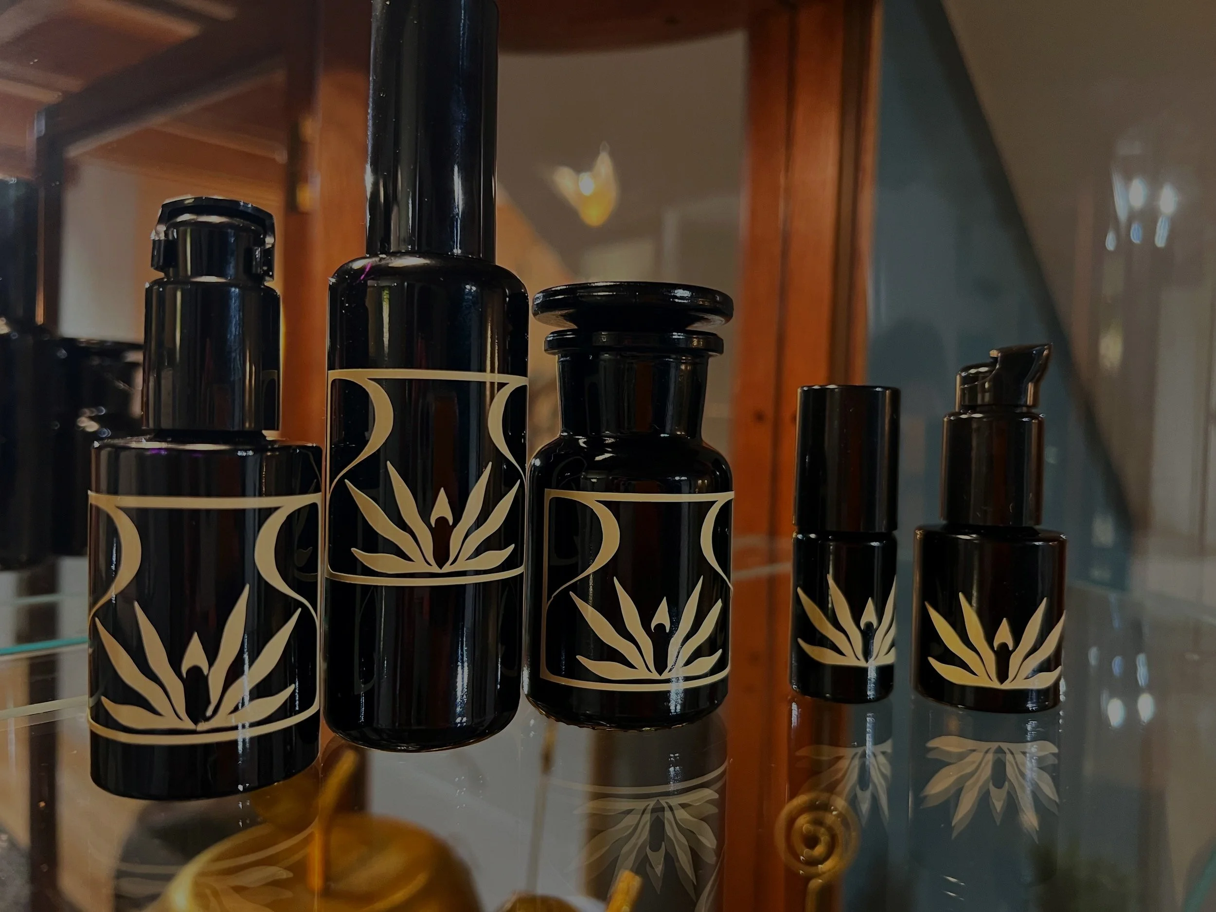 Several black bottles with cream-colored labels featuring a stylized star or plant design, displayed on a glass shelf with a wooden background.