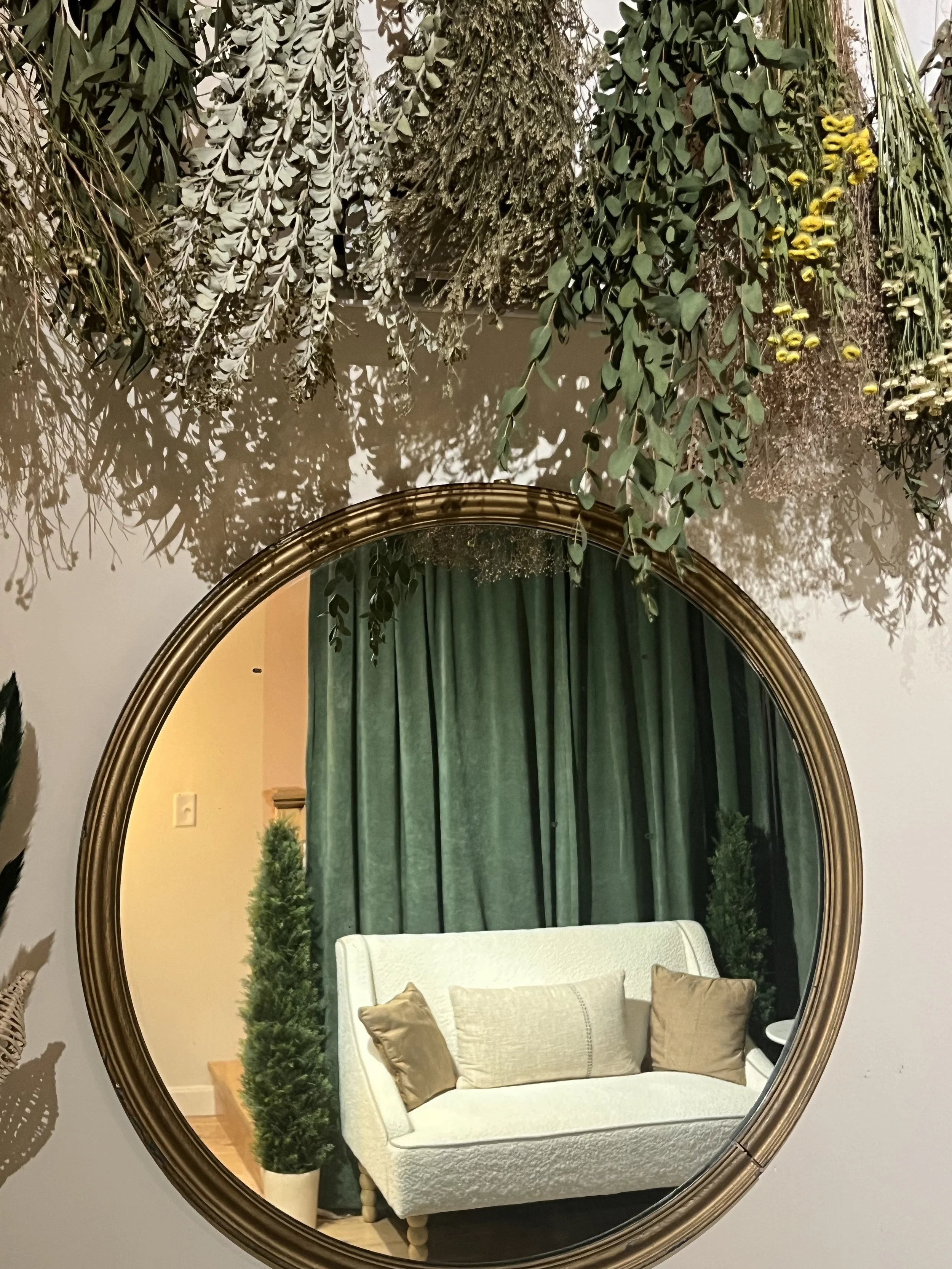 A large, round mirror with a gold frame hanging on a wall, reflecting a room with green curtains, a white upholstered sofa with two gold pillows, and two small green potted plants on either side of the sofa.