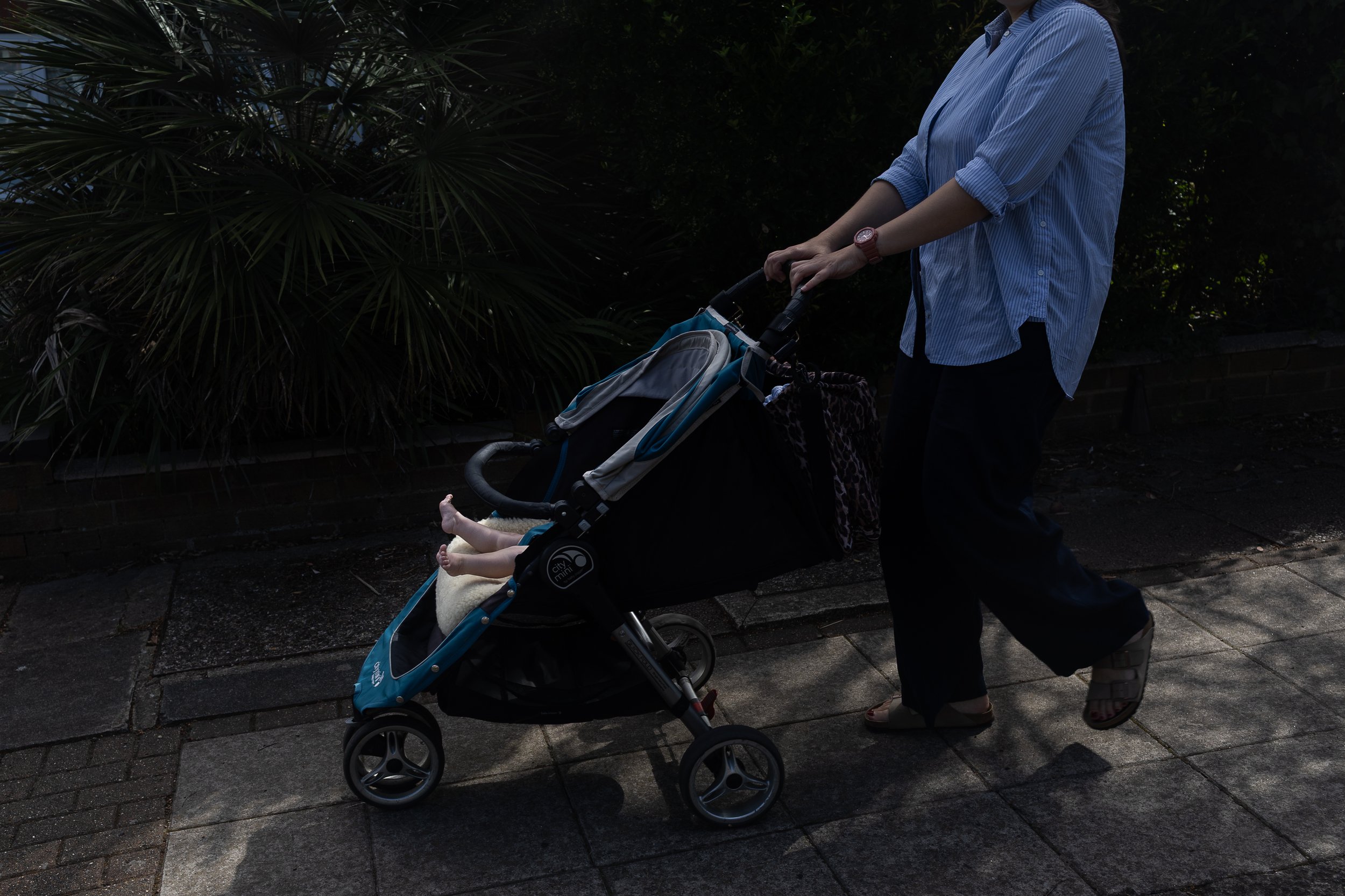 Person wearing a blue striped shirt pushing a baby stroller along a sidewalk with green shrubbery and a brick wall in the background.