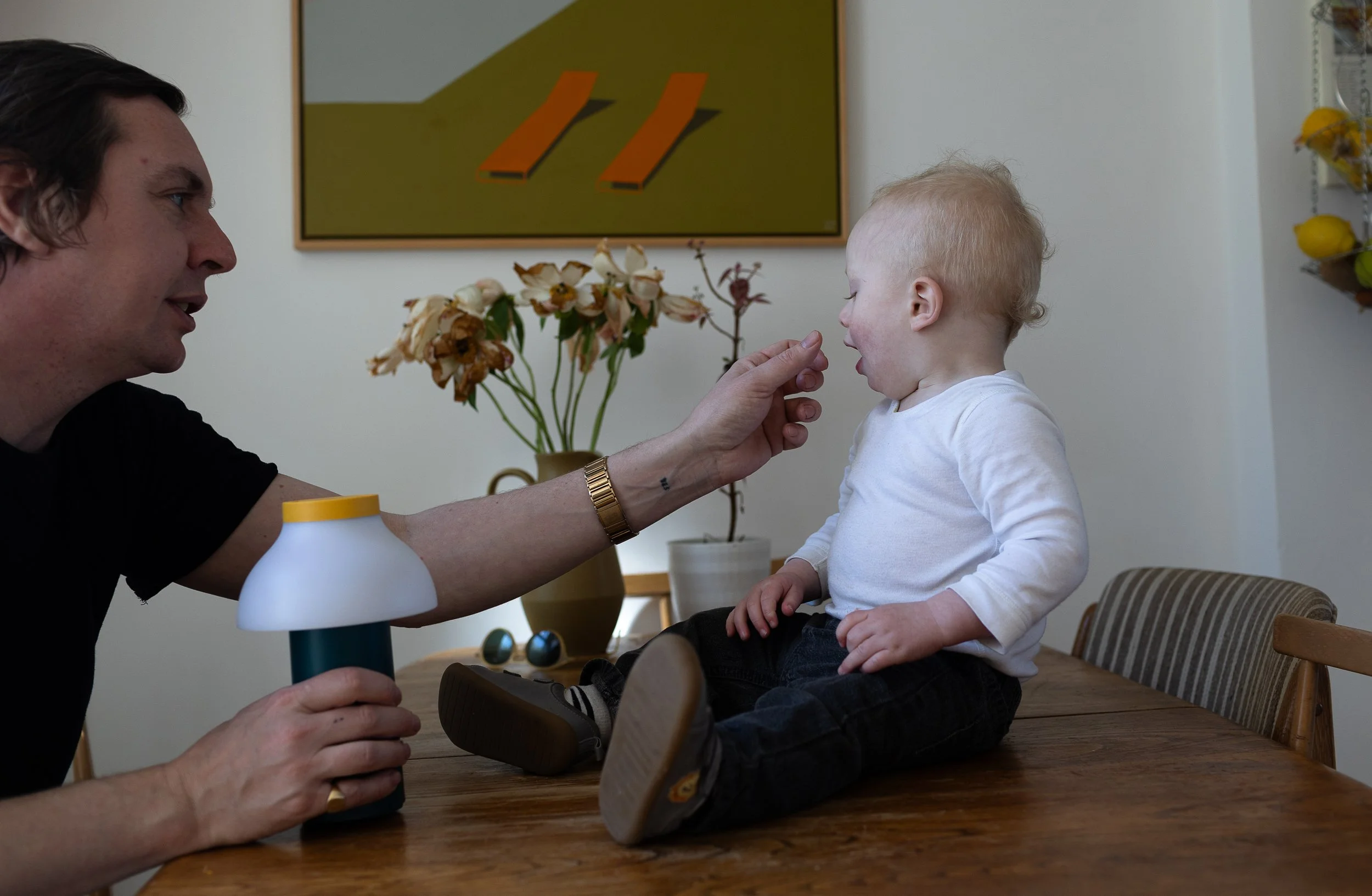A father cleans his baby sons nose as the baby sits on the table with flowers and art work in the background.