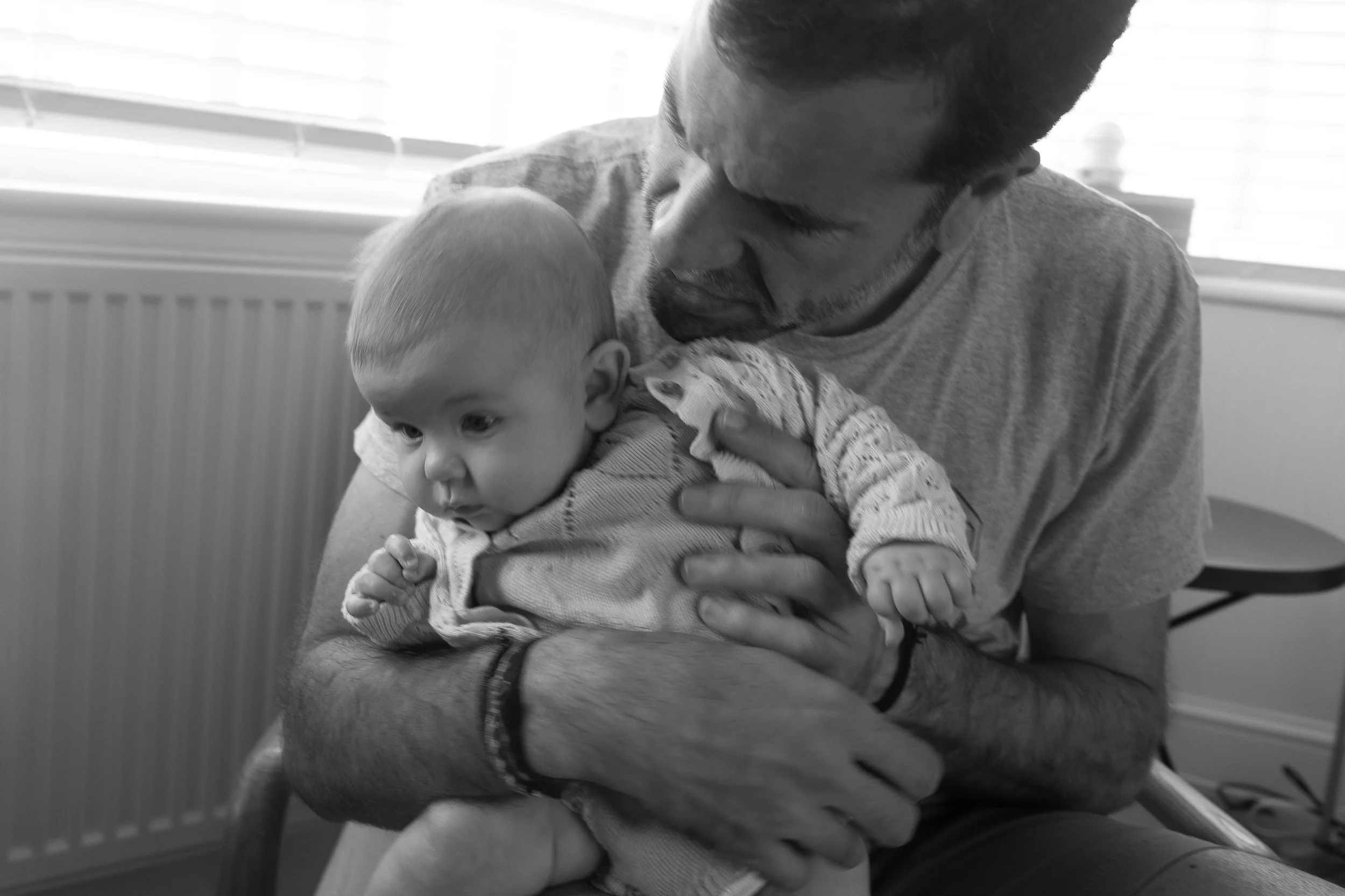 A man with a beard holds a young child with a short haircut in his arms. They are inside a room with a radiator and a window with blinds in the background. The image is black and white.
