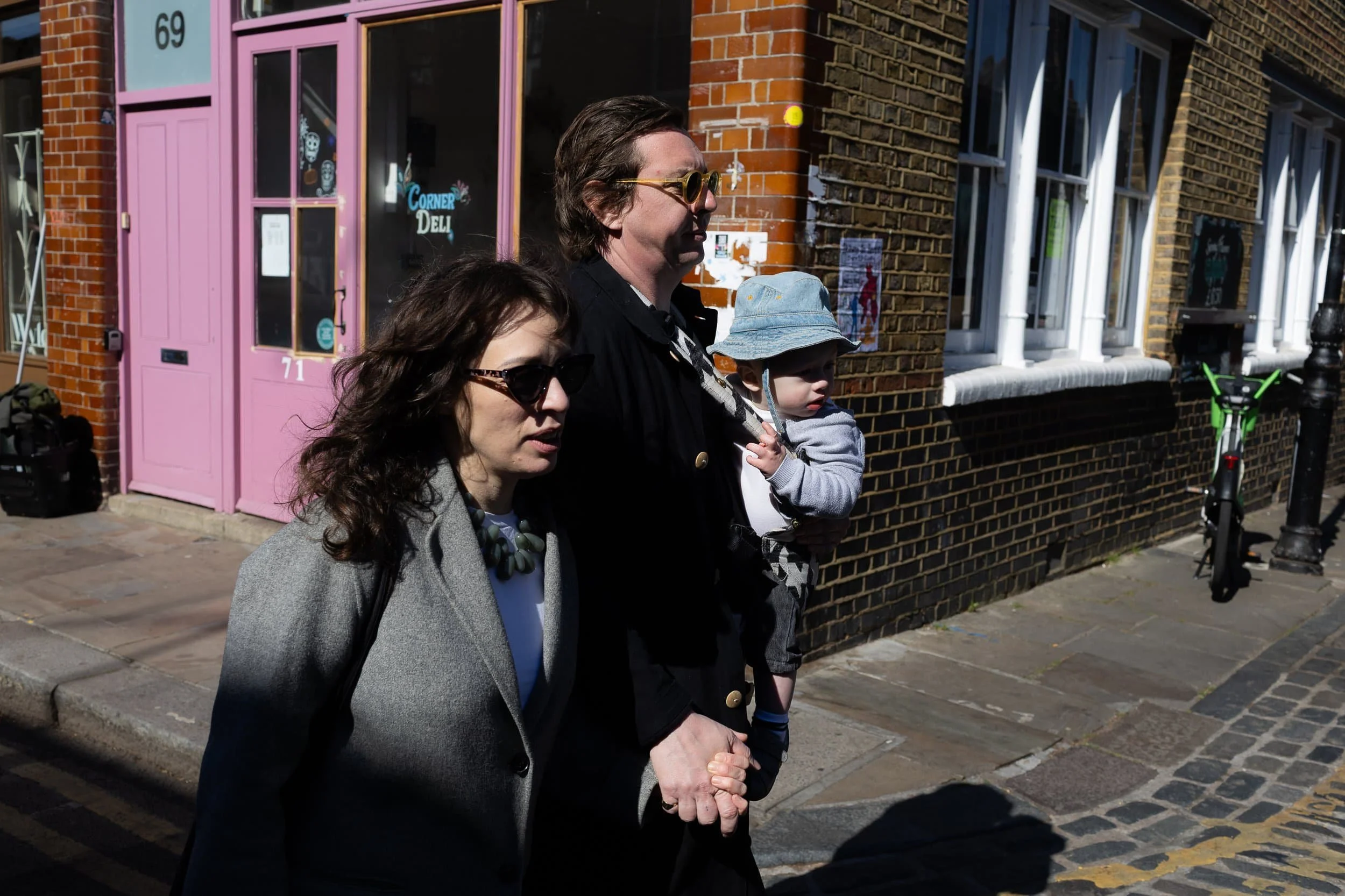 Parents walk handing hand on a sunny day in Hoxton, one carries a baby in a blue cat. Perfect for family photos.