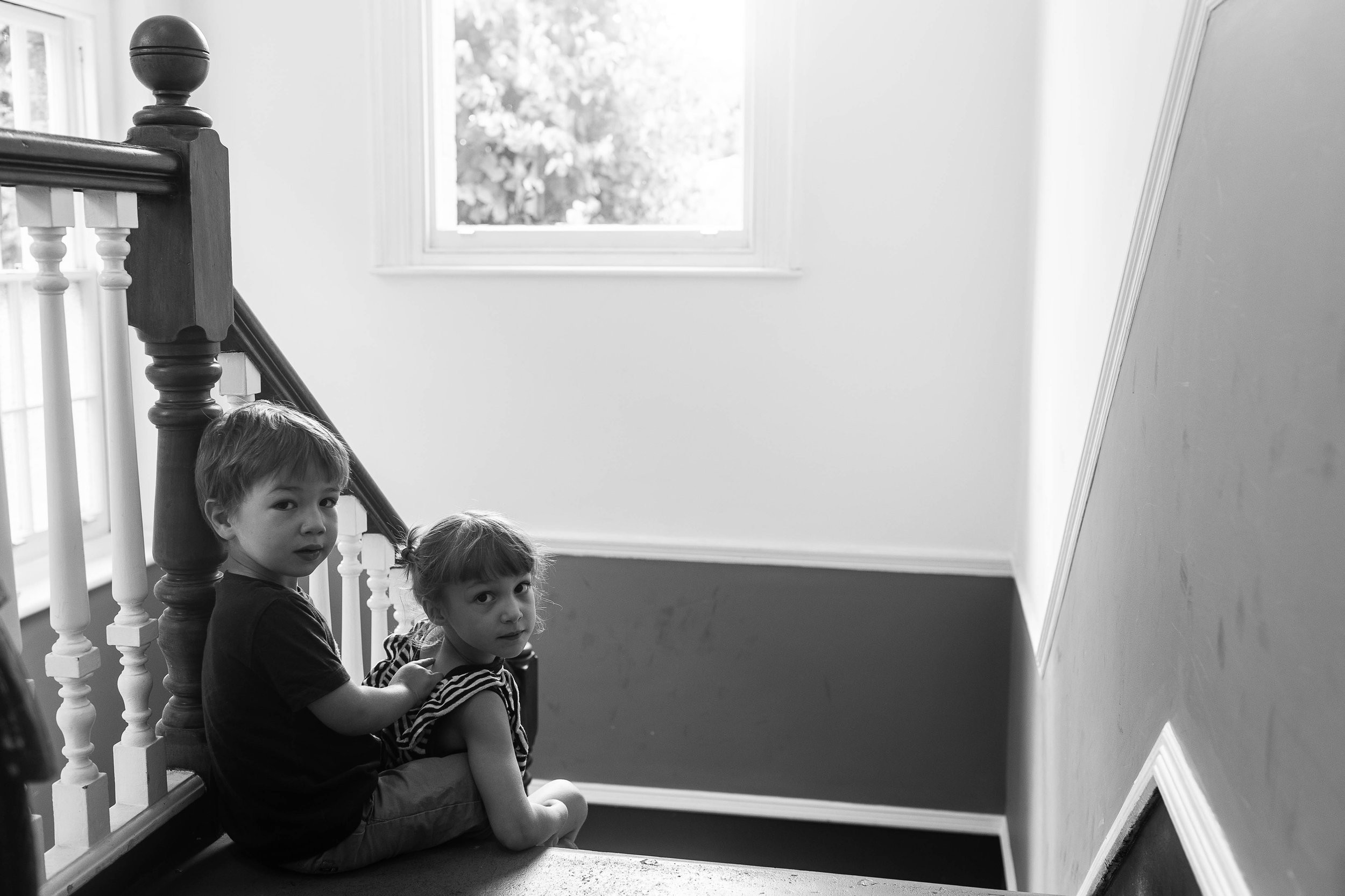 Two young children sit together on a staircase, looking at the camera in a softly lit, indoor setting.