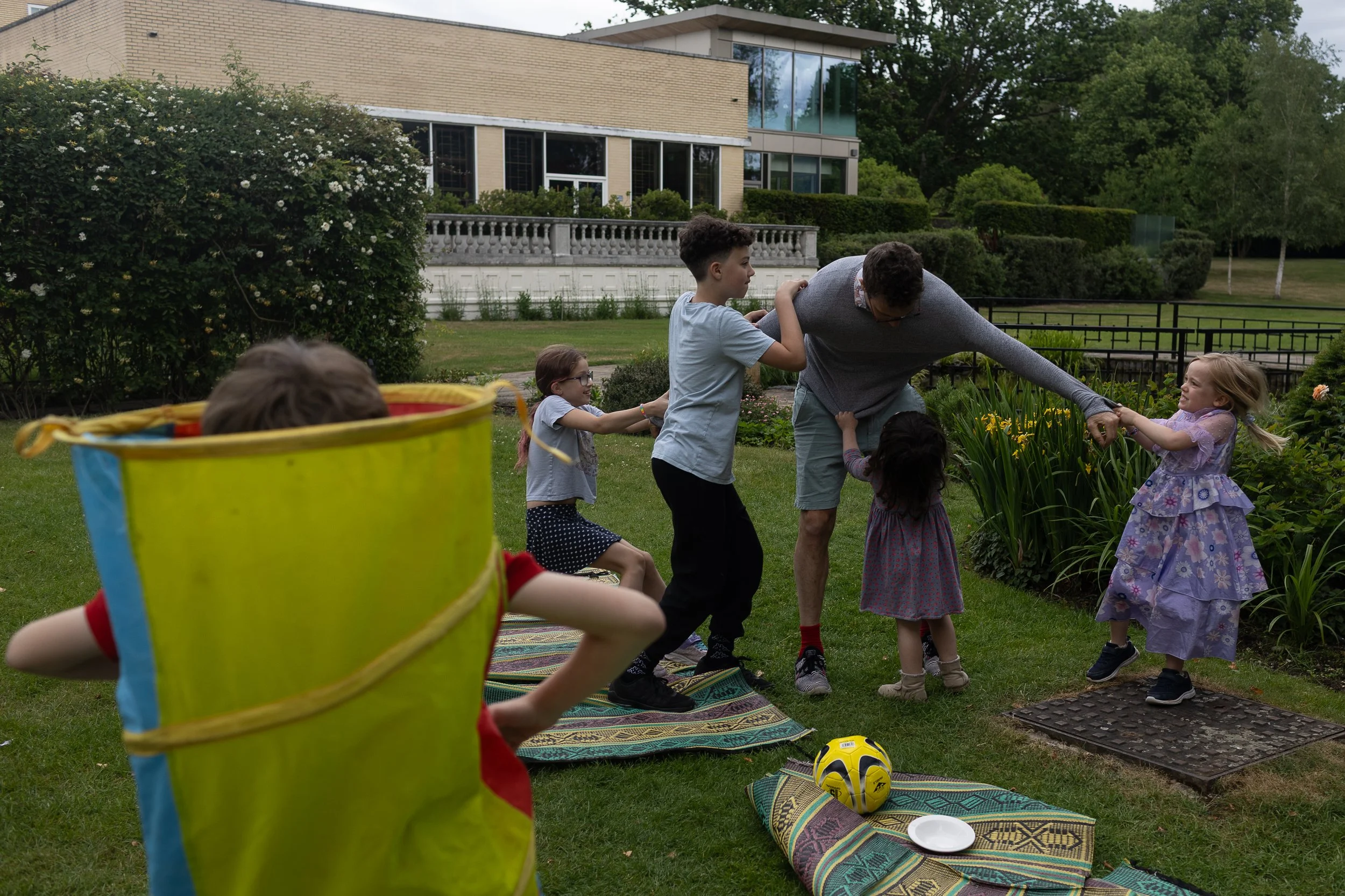 Children and an adult playing tug of war with a rope outdoors on a grassy area, with a modern house and trees in the background.