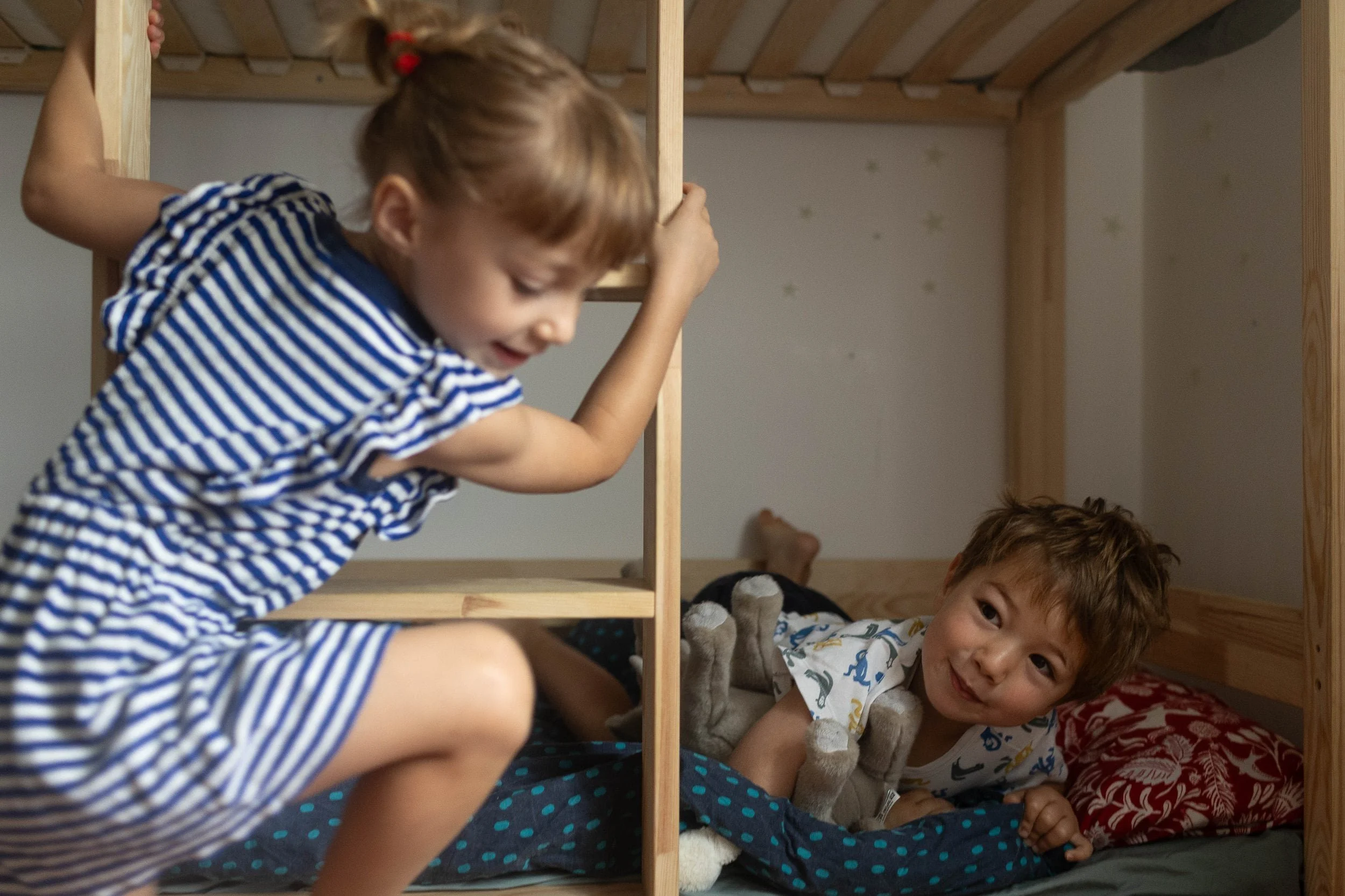 Two young children play on a wooden bunk bed; one climbs while the other lies below, smiling and holding a stuffed toy.