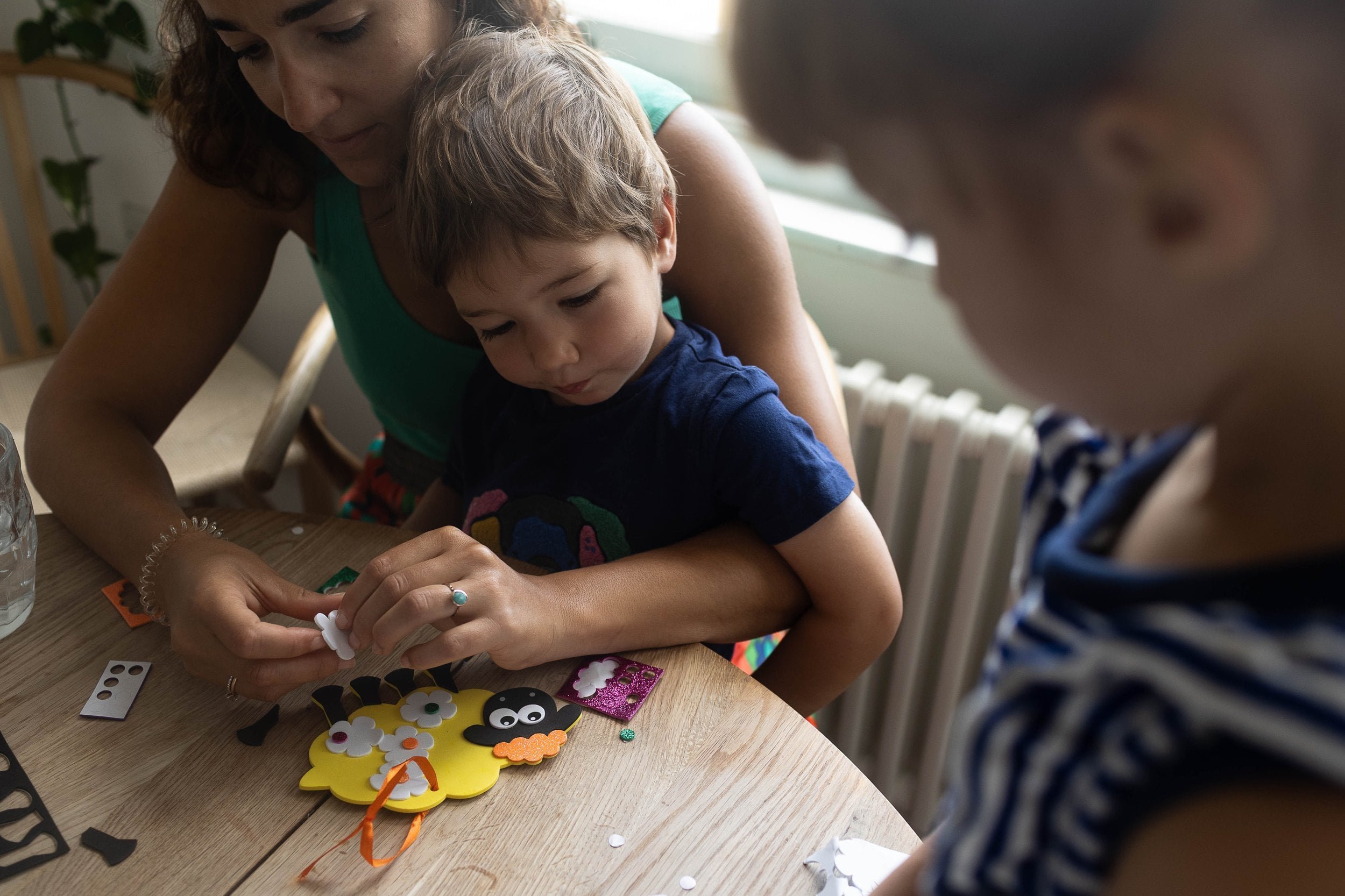 A mum helps a young boy with a craft project at a table, while another child stands nearby watching.
