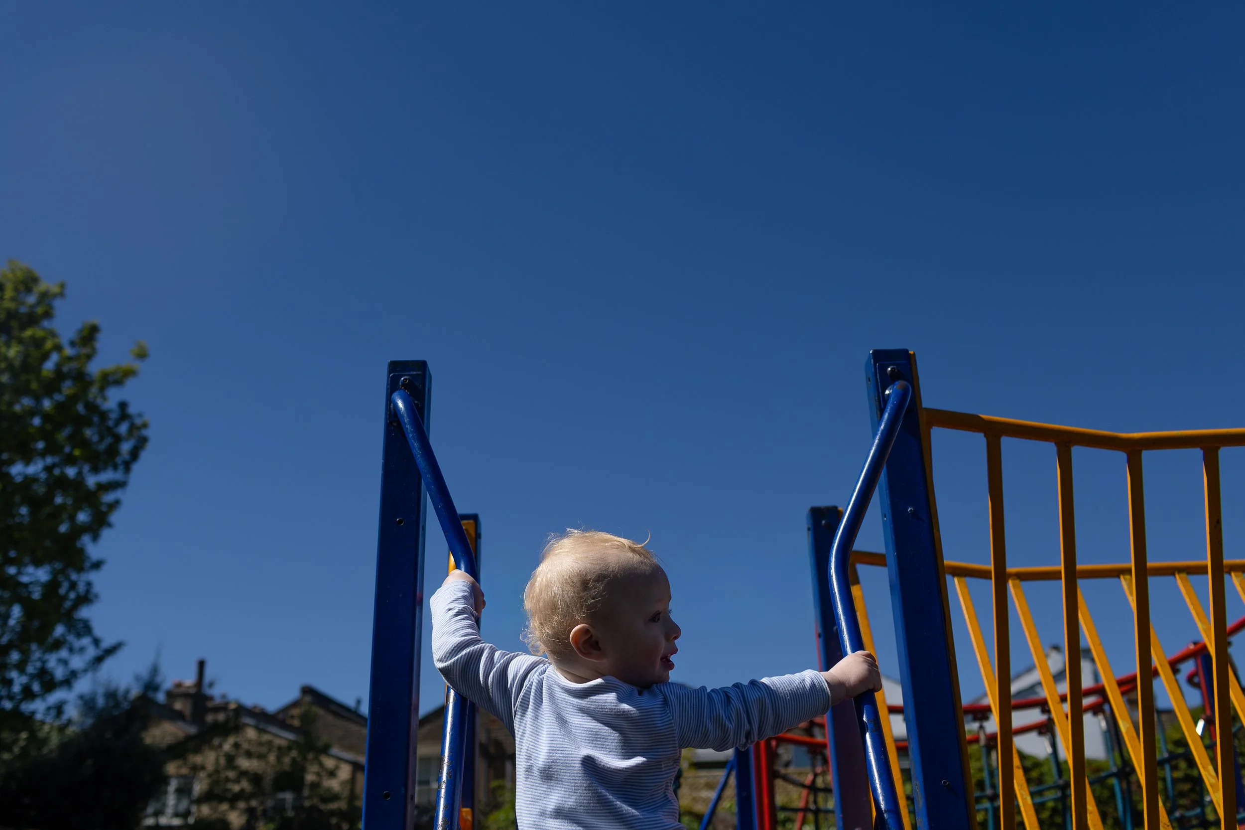 A toddler hols onto a playground bar under a clear blue sky.