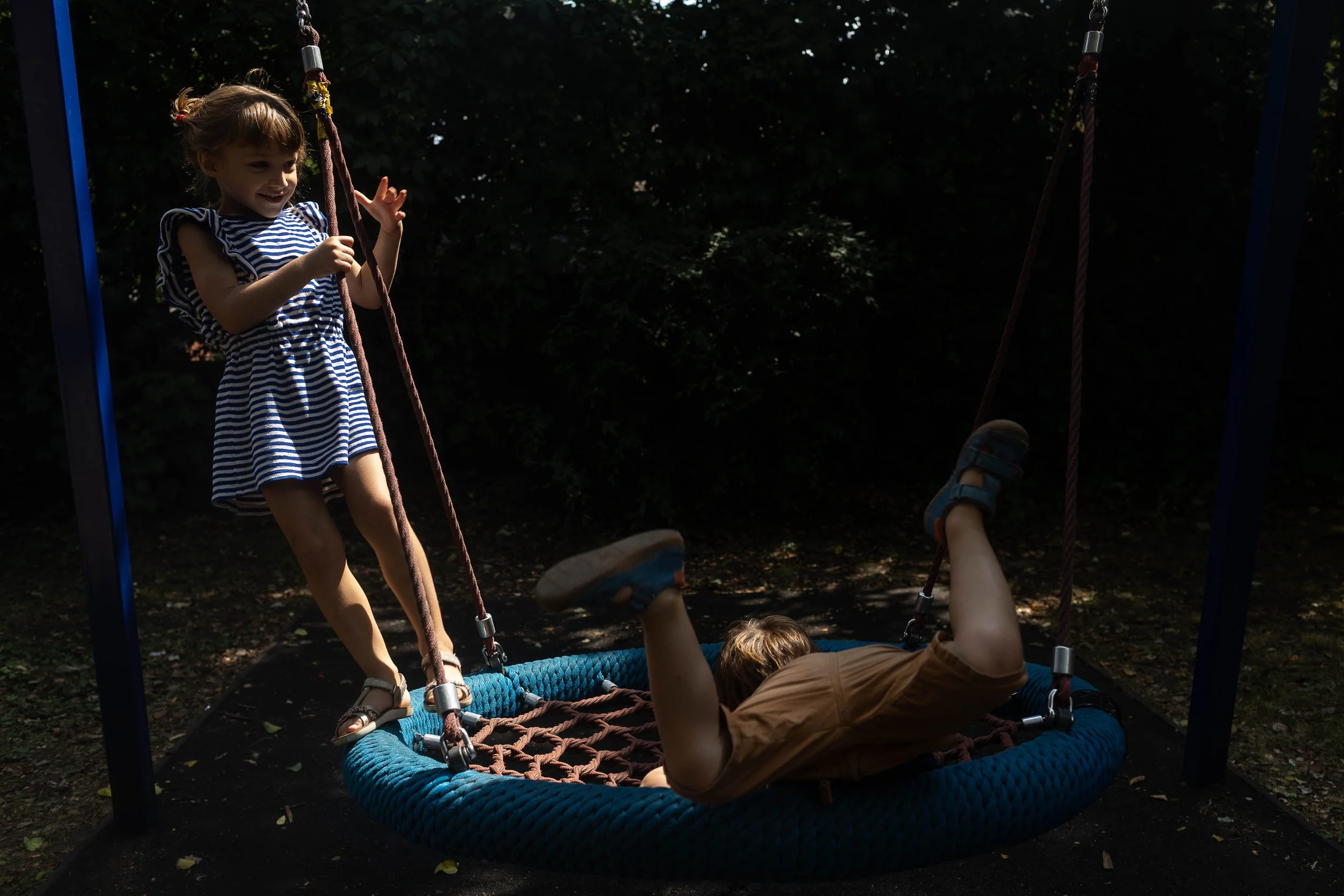 A girl in a striped dress stands by a swing while a boy lies upside down on it, both playing outdoors under shade.
