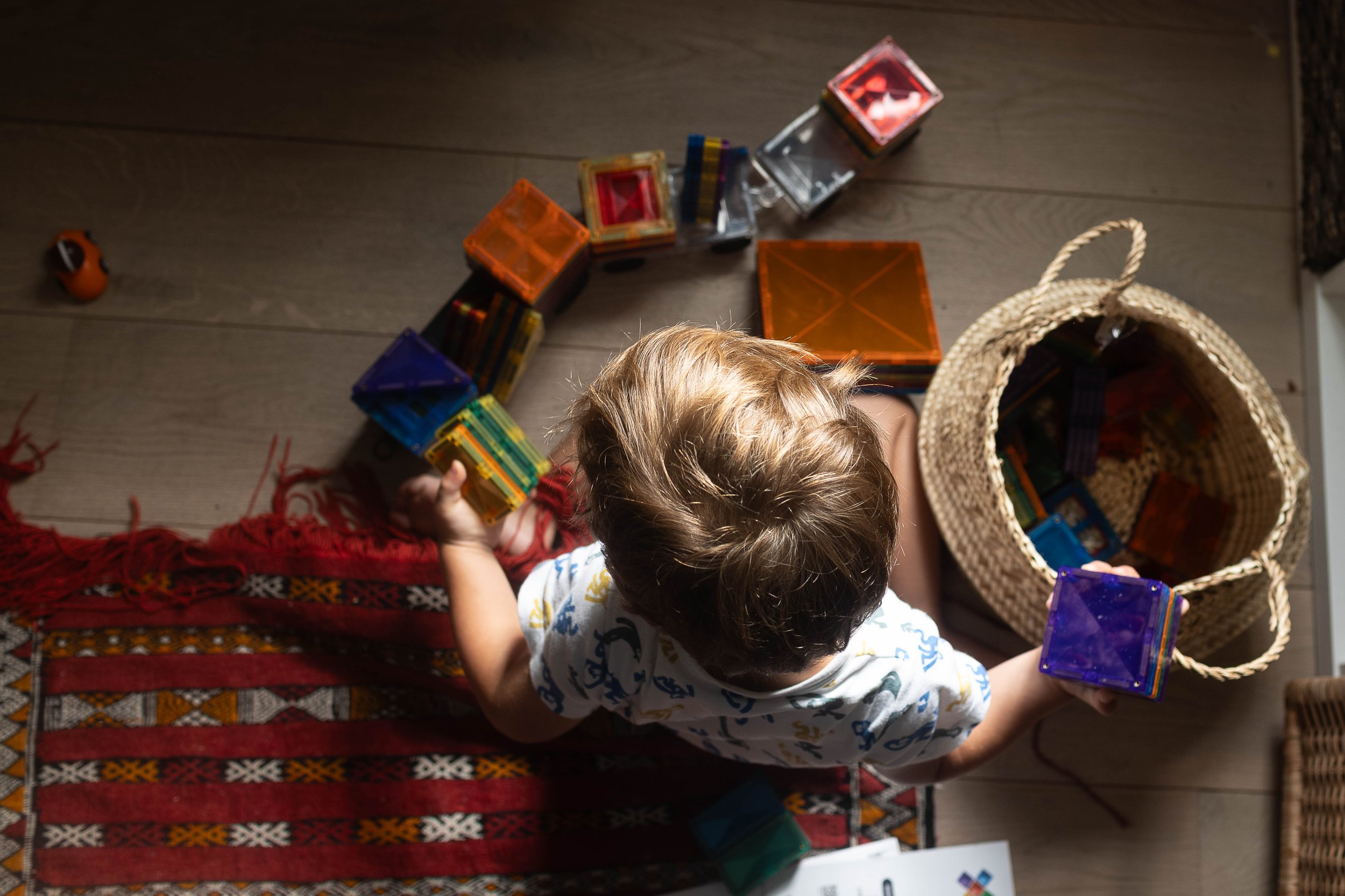 A young child sits on a rug, playing with colorful magnetic tiles next to a woven basket on a wooden floor.