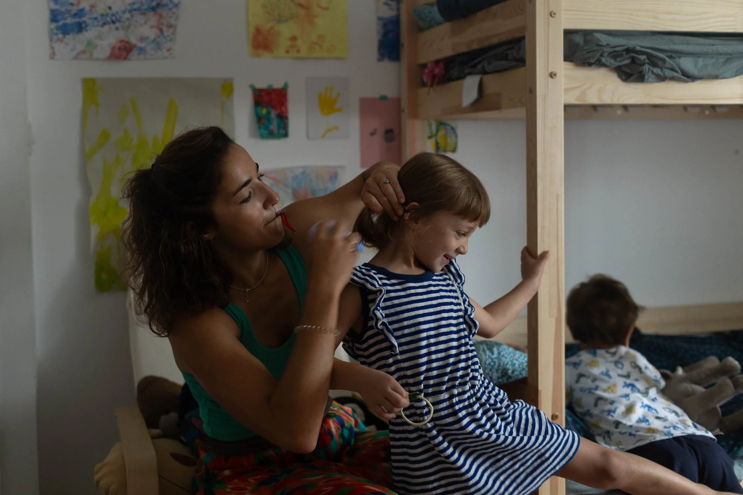 Regenerate alt text copyIconcrossIconBlue A woman brushes a young girls hair beside a bunk bed, while a boy plays on the lower bunk in a cozy, art-decorated room.