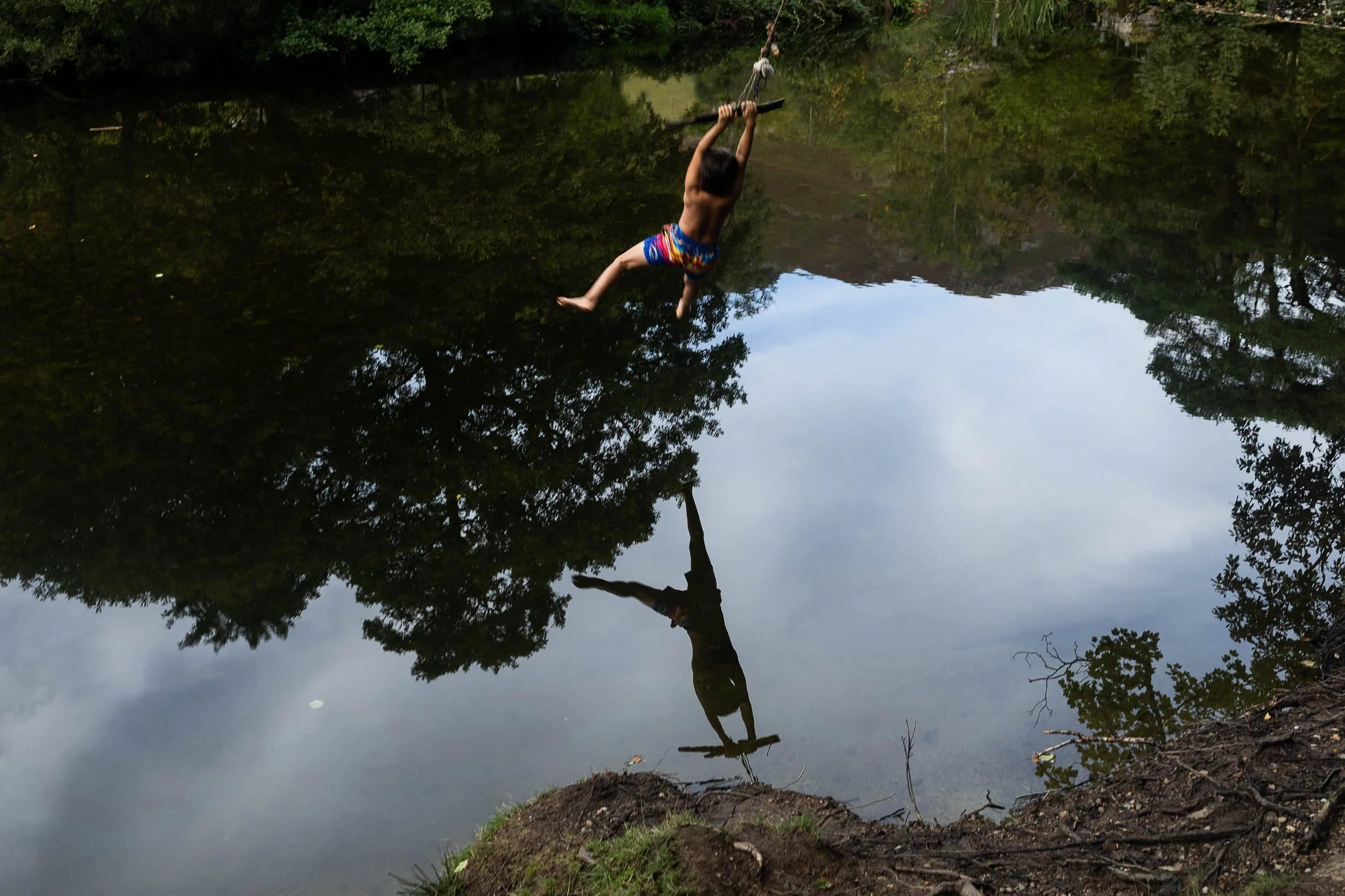 A boy swings from a rope over river reflecting trees and the child's silhouette on the water surface.