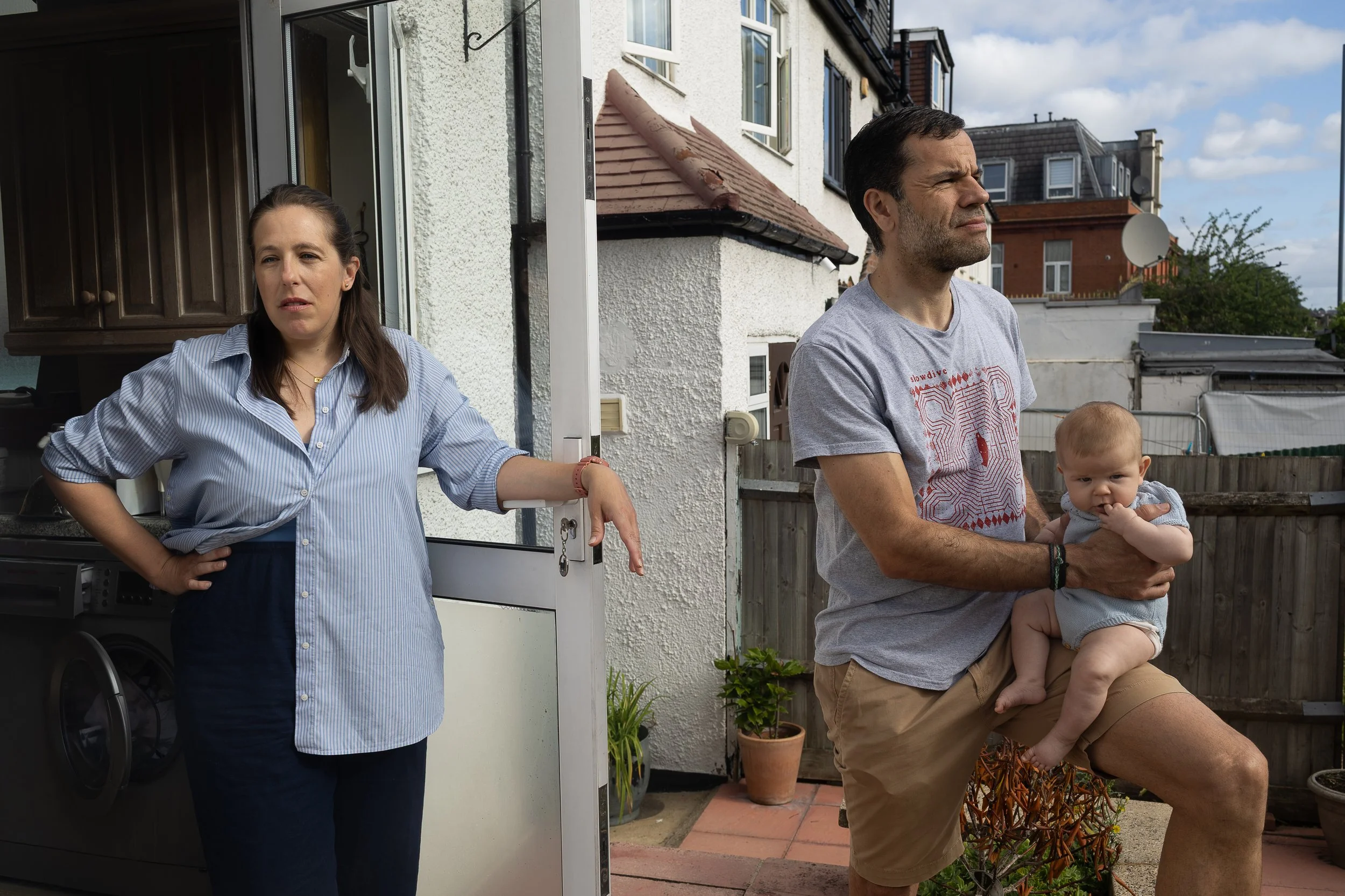A woman with long brown hair wearing a blue and white striped shirt stands outside a house, leaning on a railing, with a man holding a small child nearby. The man has dark hair, a beard, and is wearing a gray T-shirt holding a baby. The scene is set 
