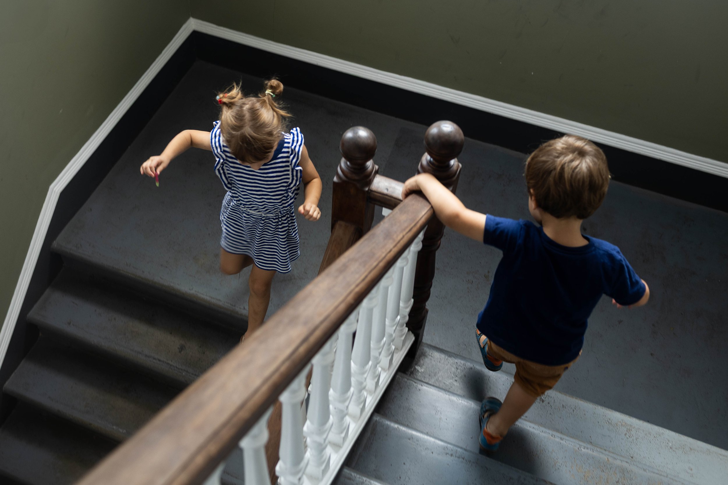 Two children, a girl and a boy, run up and down opposite sides of a staircase in a building with green walls.