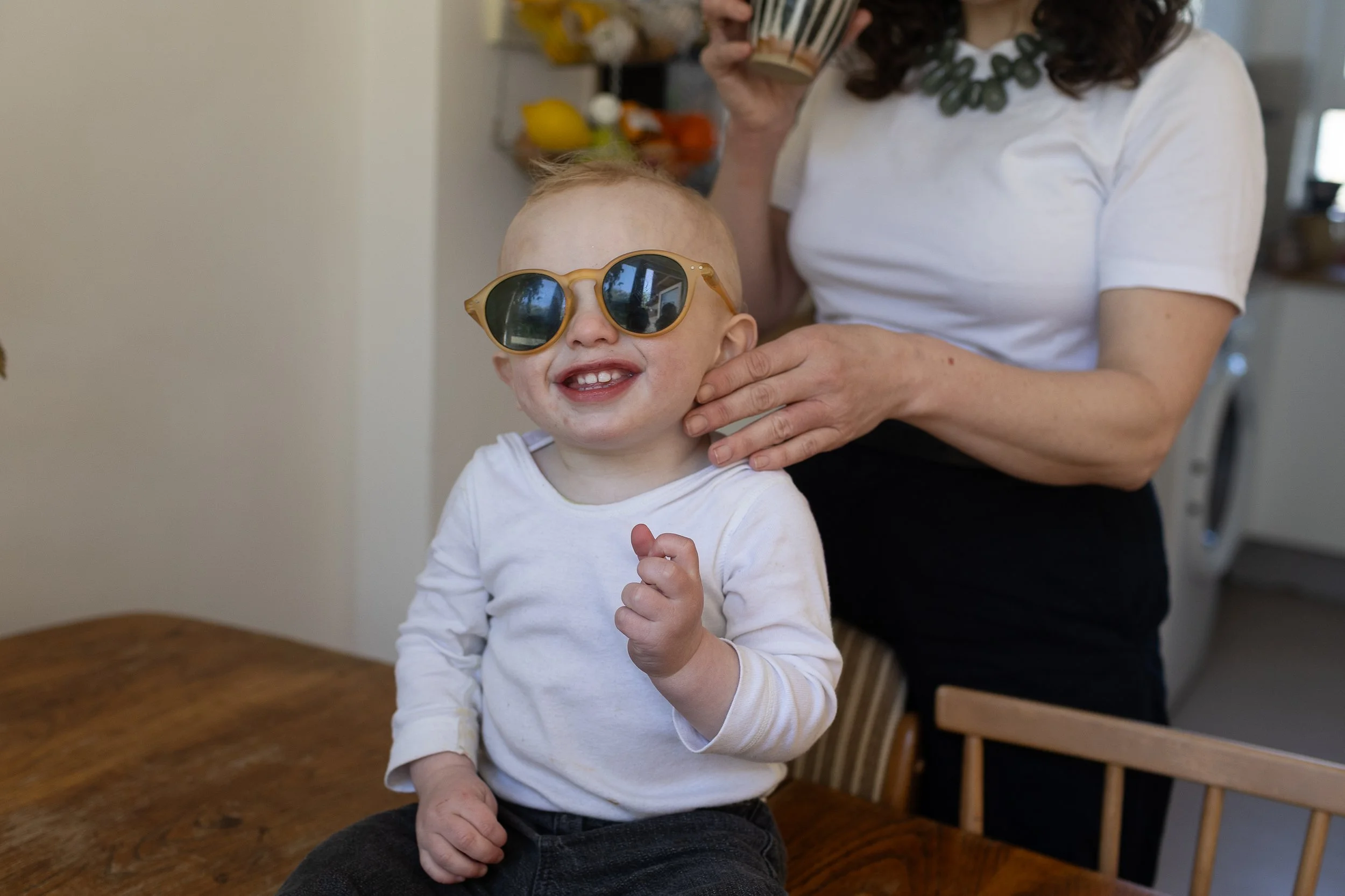A smiling toddler in sunglasses sits at a table as an adult gently touches his neck.