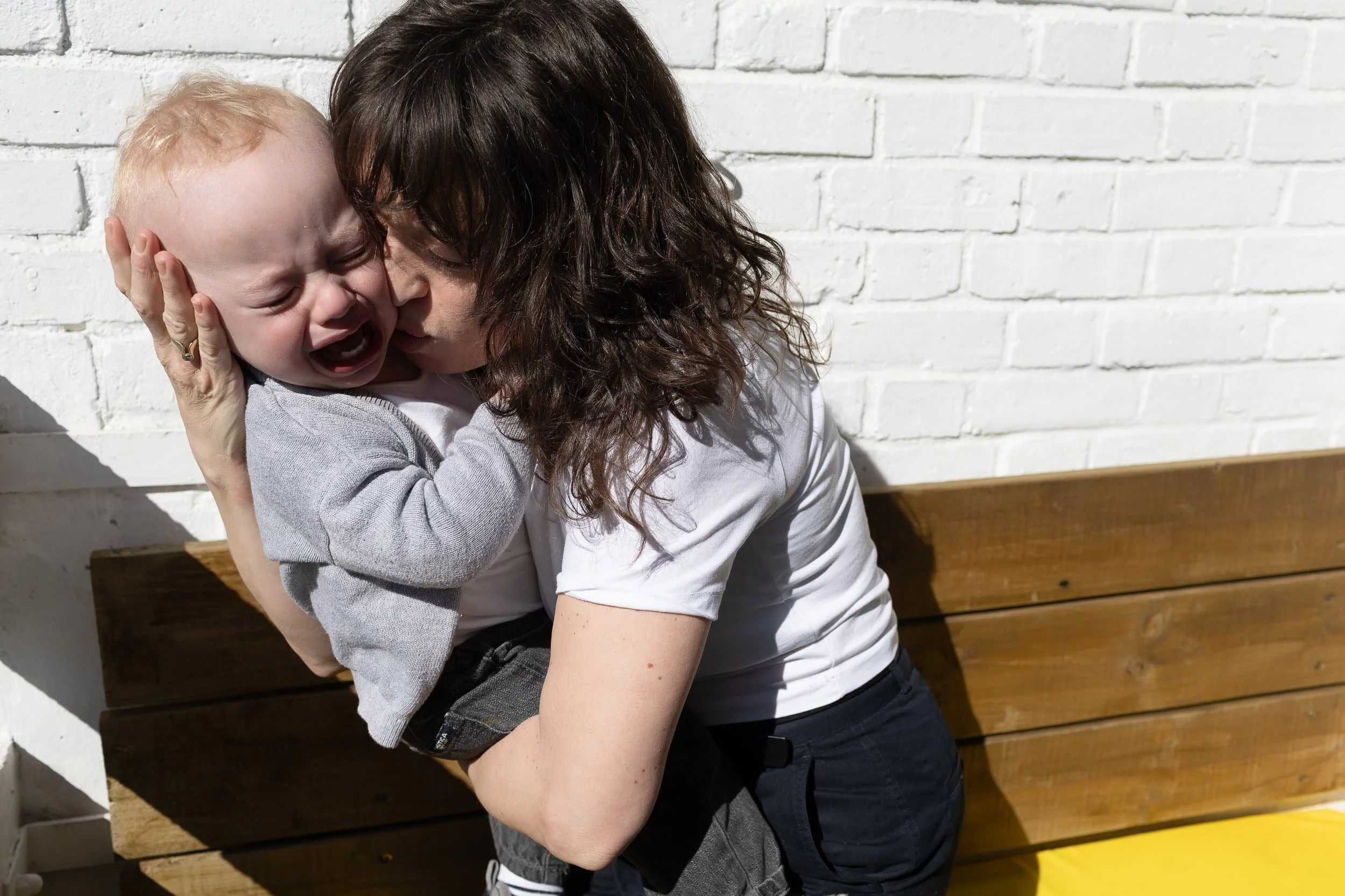 Mum comforts a crying baby outdoors, family photography in Hoxton East London on a sunny day against a white brick wall.