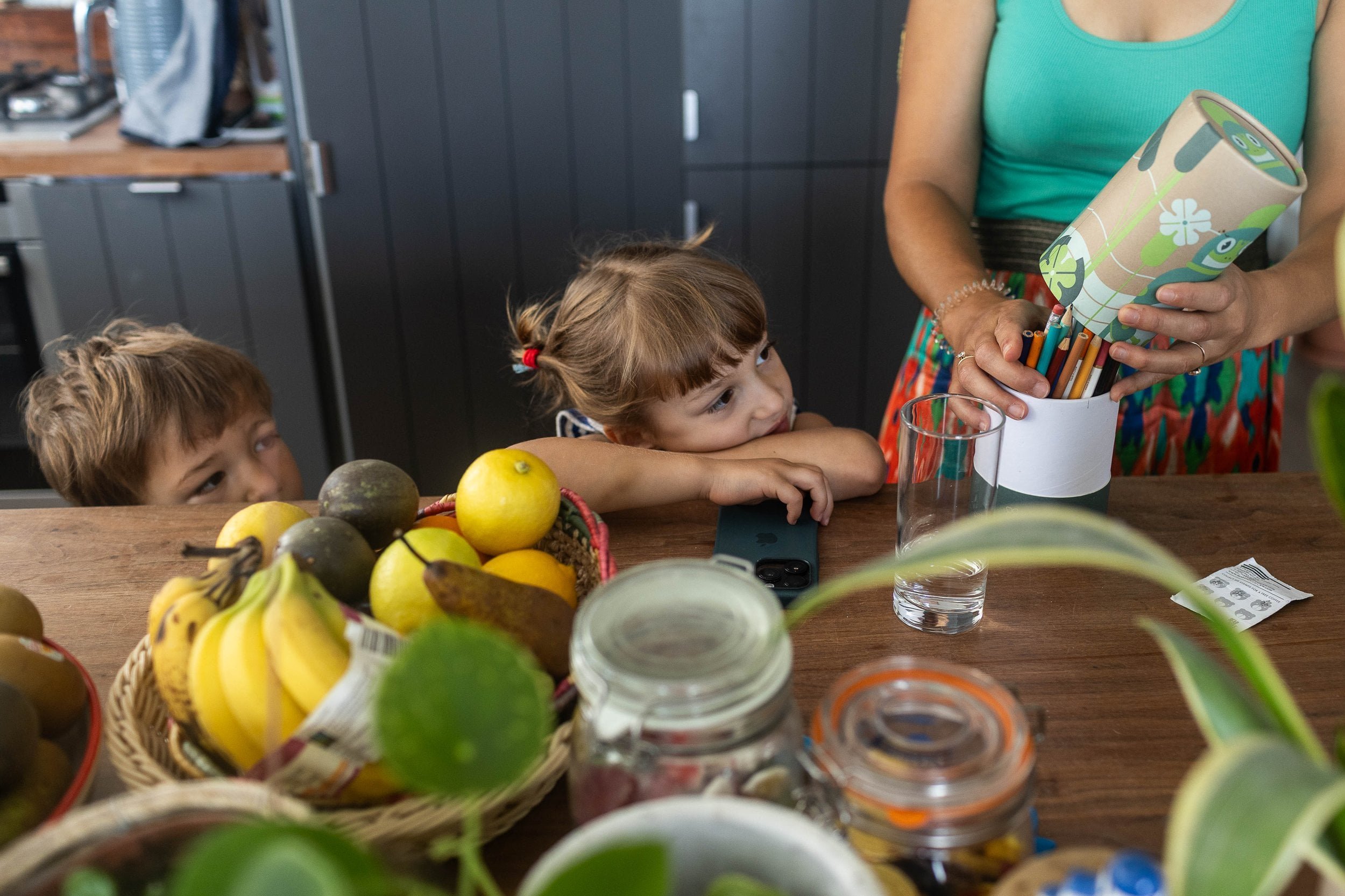 Two children watch a woman place colored pencils in a cup on a kitchen counter with fruit and jars.