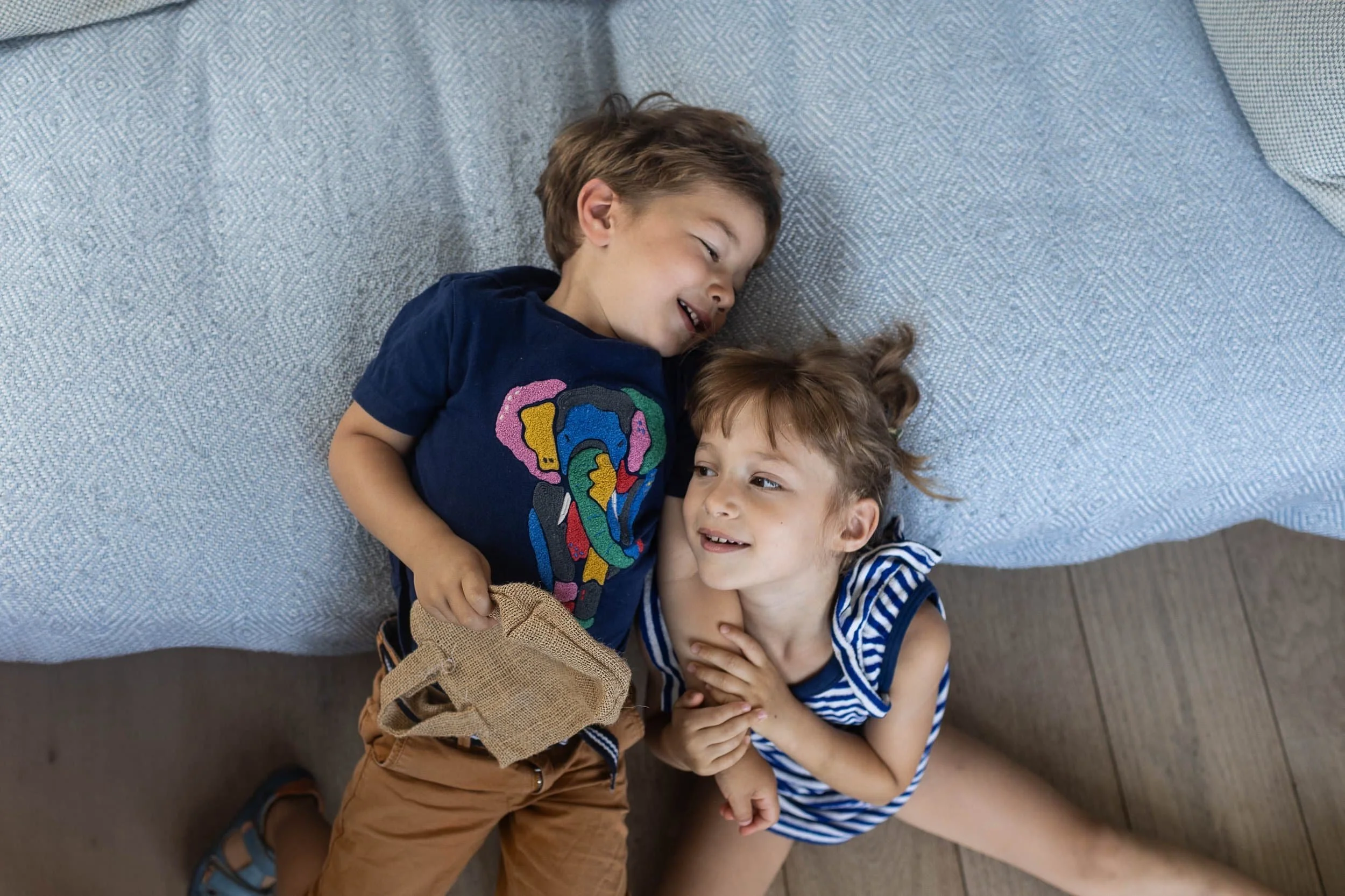 Two young children, a boy and a girl, lying and smiling together on the floor next to a blue sofa.
