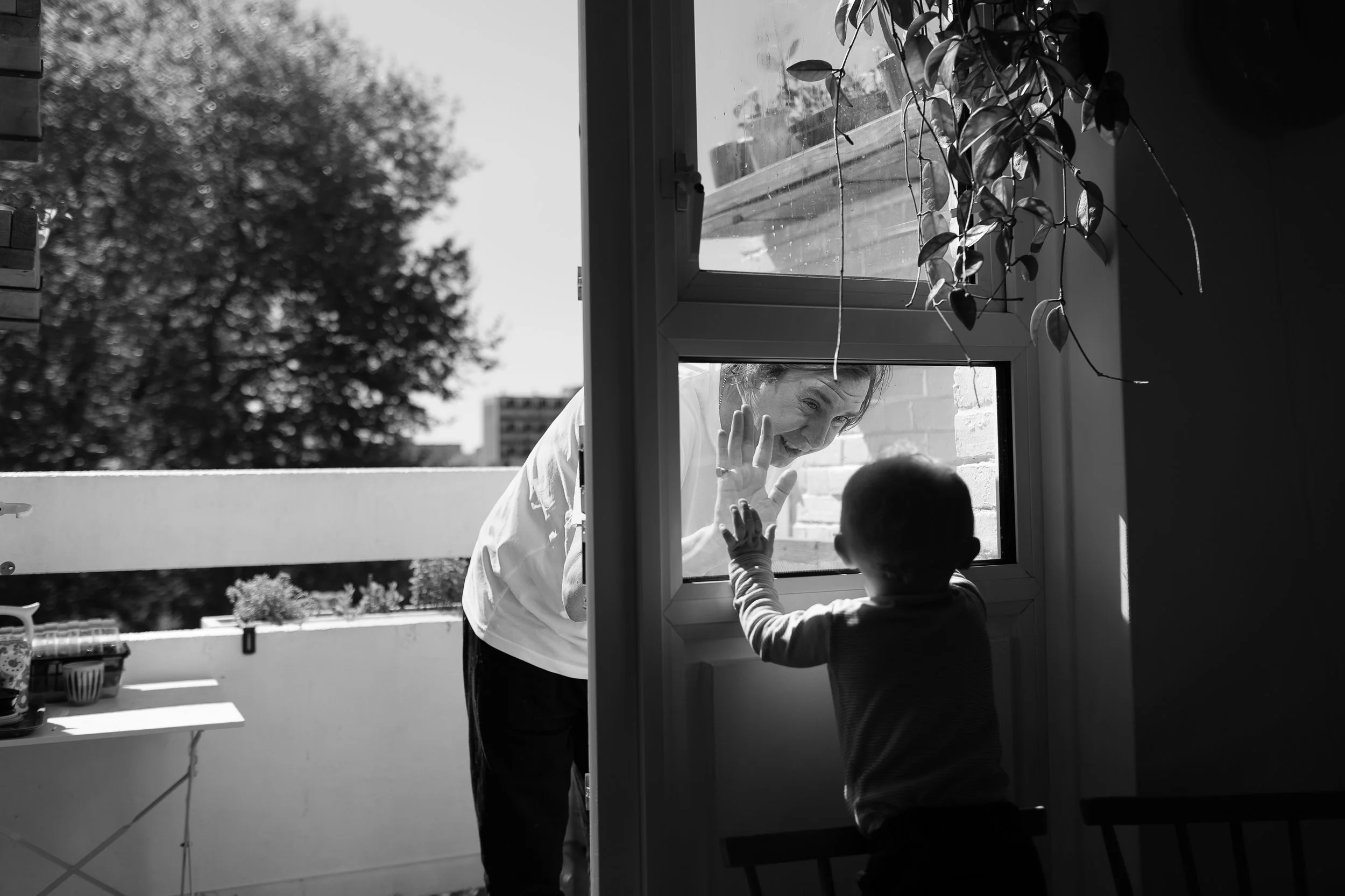 A child touches a glass window, reaching towards an adult smiling outside - a timeless moment by a family photographer in Hoxton
