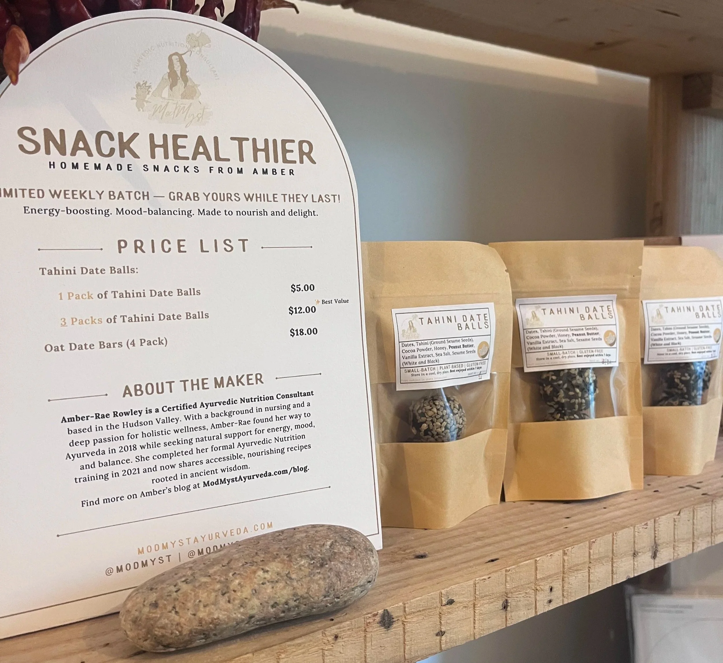 A display of homemade tahini date balls in paper bags on a wooden shelf, with a menu card describing prices and information about the maker, Amber-Rae Rowley, an Ayurvedic nutrition consultant.