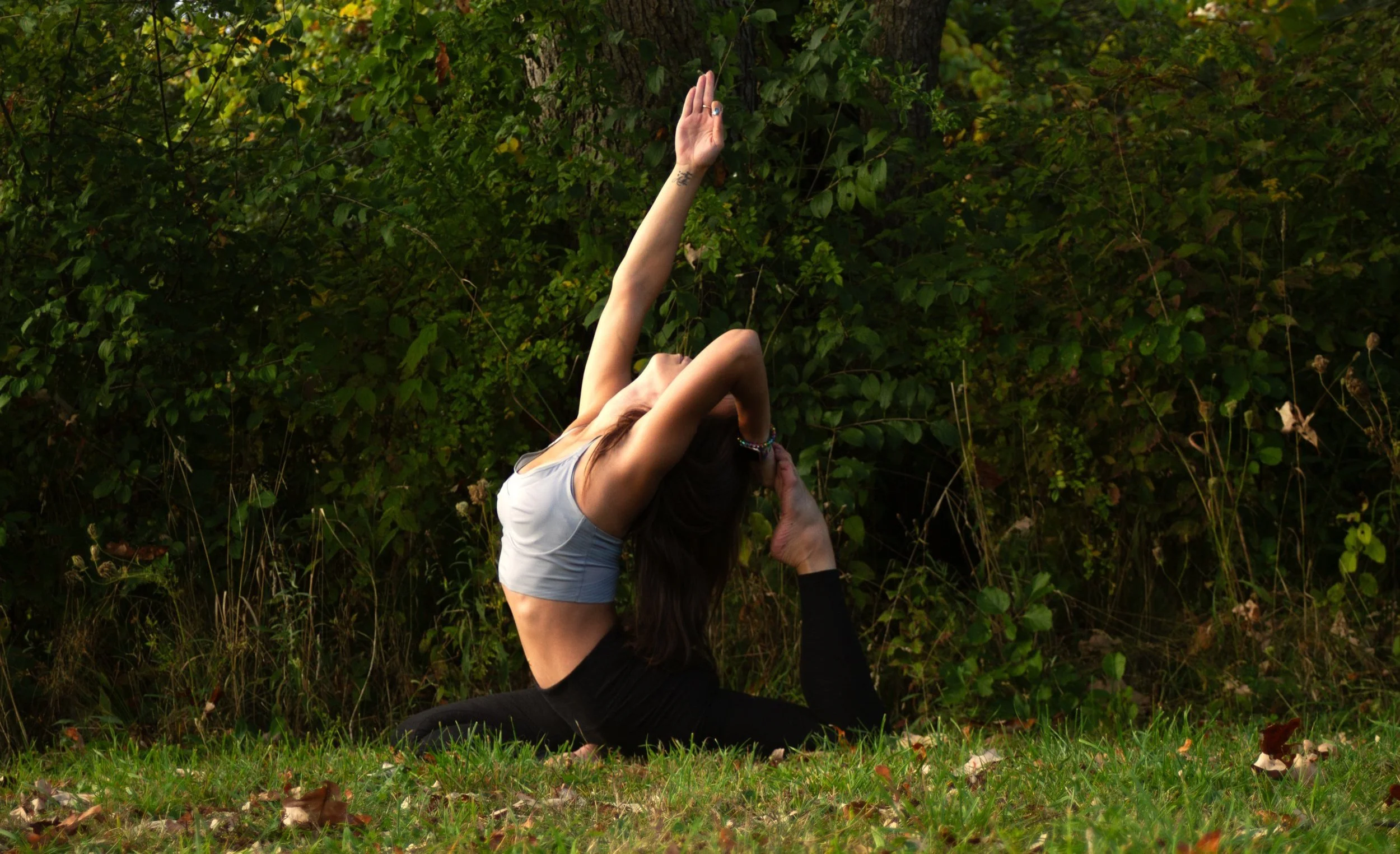 Woman practicing yoga outdoors, performing a backbend stretch on grassy ground in front of green bushes.