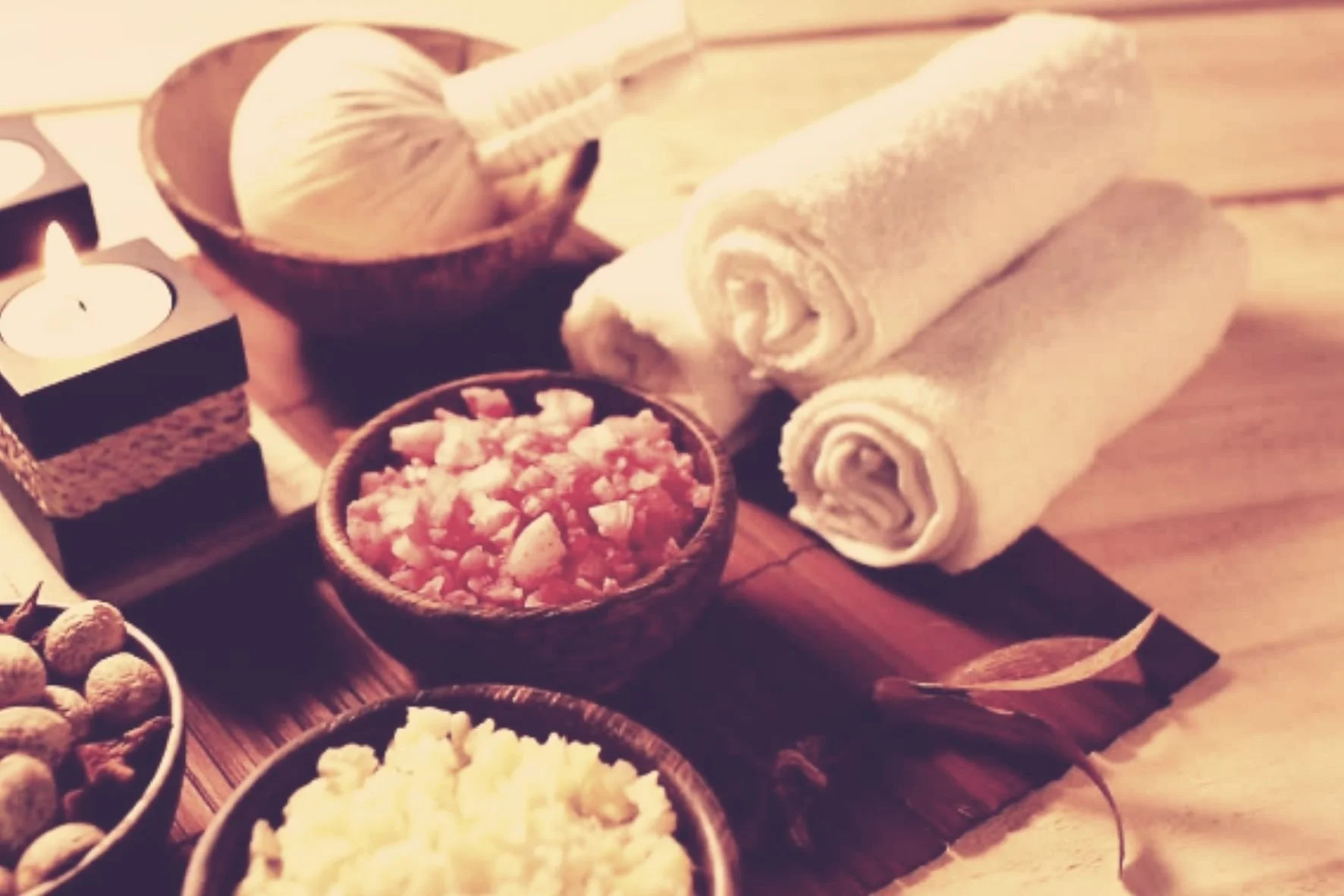 Spa setting with rolled white towels, bowls of scrub, and a lit candle on a wooden tray.