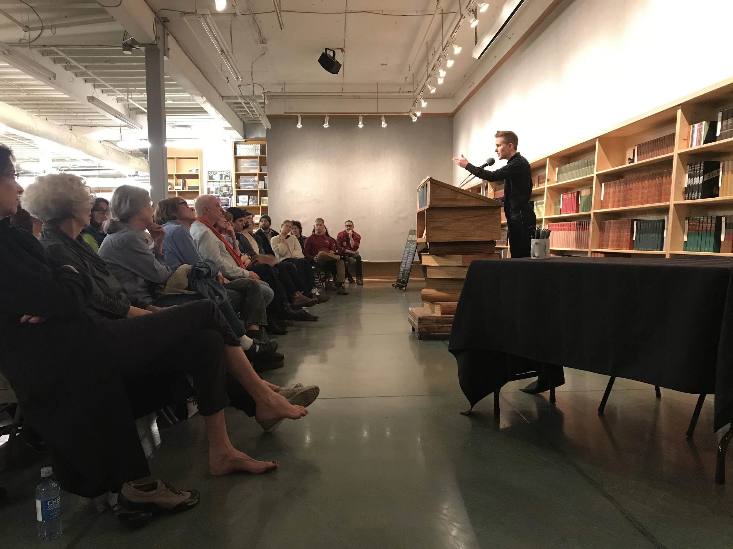 Sandy Allen speaking at Powell's in Portland in 2018 before a packed house. He's behind a podium and there's someone barefoot in the first row.